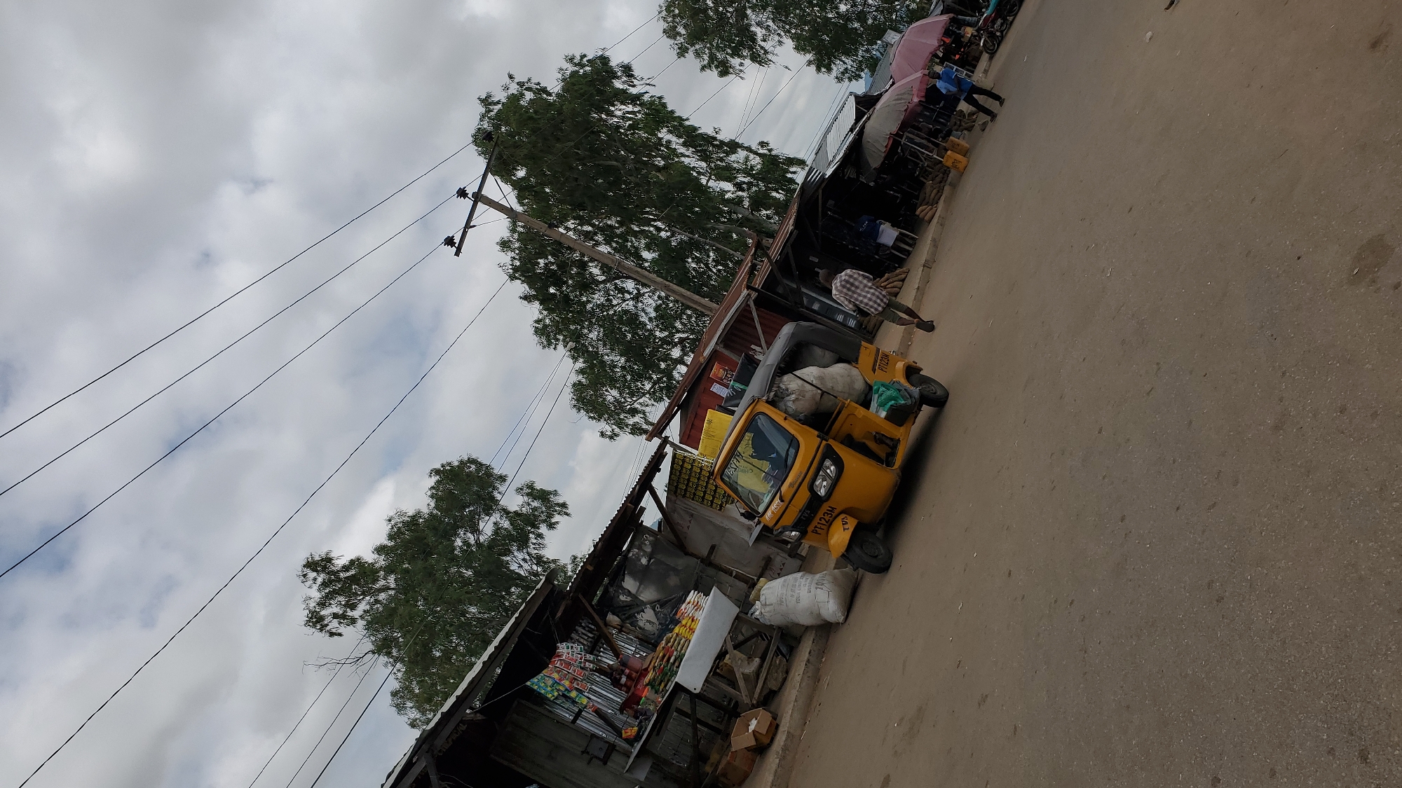 The image depicts a street scene in Jalingo, Nigeria, likely a market area. In the foreground, a yellow auto-rickshaw, possibly a commercial vehicle, is parked on an asphalt road. The auto-rickshaw is loaded with several large white sacks. To the left of the auto-rickshaw is a roadside stall or shop with a corrugated metal roof, displaying various goods such as packaged food items and produce. Several large white sacks are also placed on the ground in front of this stall. A person in a plaid shirt and shorts is walking away from the camera in the middle ground, heading towards other stalls in the background. Further back, more market stalls with awnings are visible, some displaying umbrellas. Power lines are strung across the sky, supported by utility poles. Trees with dense foliage are scattered throughout the scene, indicating a somewhat rural or semi-urban environment. The sky is overcast with clouds. The overall impression is a bustling, functional street market setting.