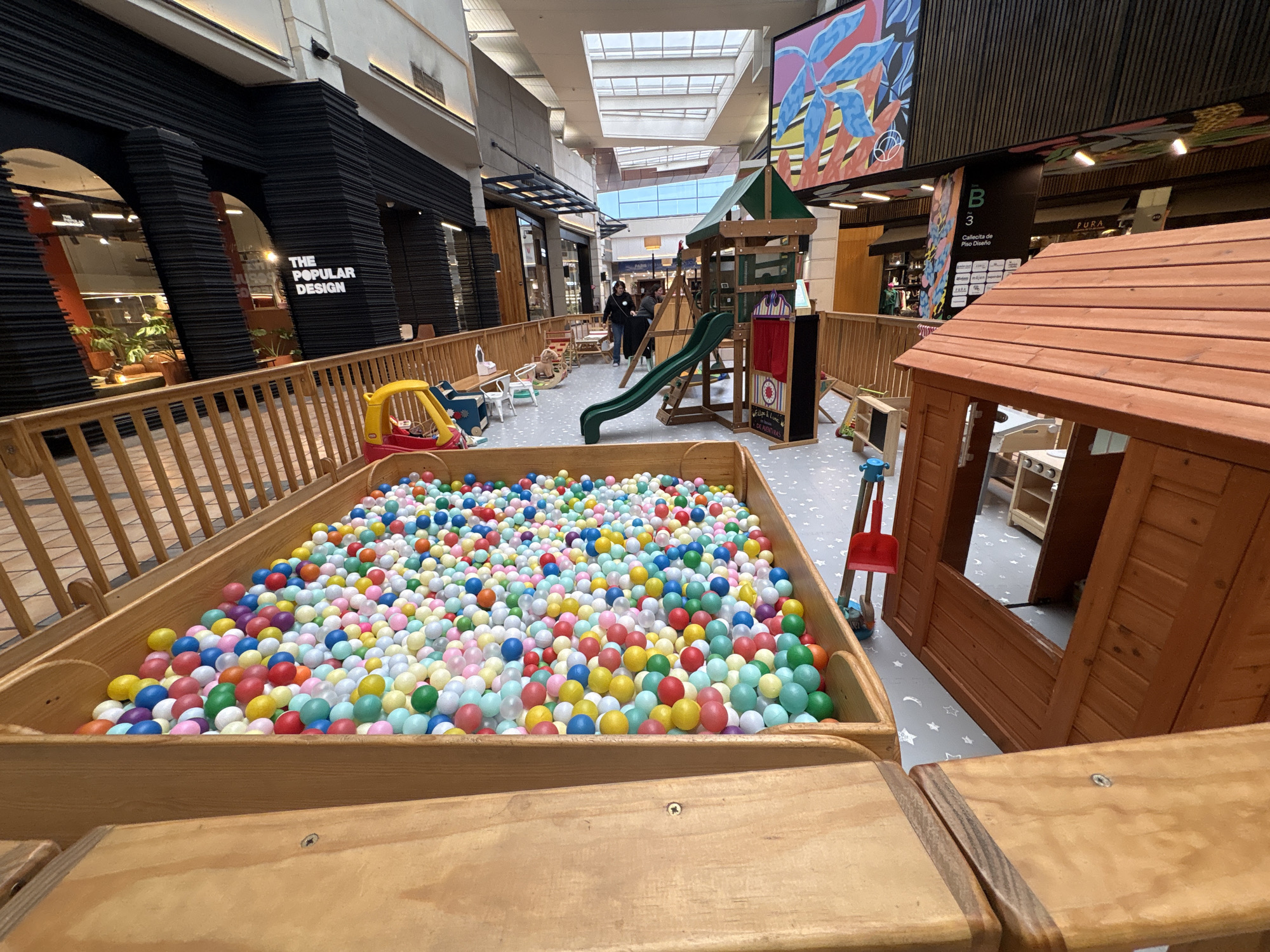 The image shows a brightly lit indoor children's play area, likely located in a shopping mall in Santiago, Chile. The foreground is dominated by a large wooden ball pit filled with a multitude of colorful plastic balls in shades of red, yellow, blue, green, pink, and white. To the left, a section of a wooden fence is visible.
In the mid-ground, a green plastic slide descends into the play area. Adjacent to the slide is a wooden play structure that resembles a small fort or castle, complete with windows and a roof. A small red toy shovel rests near the base of the play structure. Further left, there is a yellow toy car and some small white chairs and tables. Two people, a woman and a child, are visible near the play structure.
The background reveals the architectural features of a modern indoor space, likely a mall. Large windows and skylights allow natural light to filter in. On the left side of the image, a storefront with the text "THE POPULAR DESIGN" is visible, indicating a retail establishment. A large, colorful mural depicting abstract floral elements adorns the wall in the upper right portion of the image. Signage with "B 3" and "Callecita de Piso Diseño" suggests a specific section or floor within the mall. The overall environment is clean and well-maintained, designed for children's recreation.
