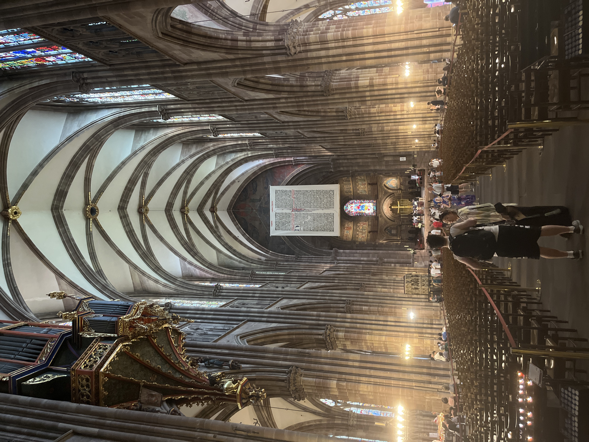 This media file captures an interior view of a grand cathedral, likely during daylight hours given the illumination. The architecture is Gothic, characterized by soaring vaulted ceilings, intricate stone ribbing, and tall, arched windows that feature stained glass.
Prominent objects within the scene include a large, ornate pipe organ on the left, with dark pipes and gilded decorative elements. In the center, a large, open book, possibly a Bible or liturgical text, is displayed. Rows of dark wooden pews fill much of the floor space, leading towards the altar area at the far end. Several people are visible, dispersed throughout the cathedral. Some are seated in the pews, while others are standing or walking, appearing to be tourists or worshippers. The overall atmosphere seems peaceful and reverent, with the vastness of the space and the architectural grandeur creating a sense of awe. The lighting suggests it is daytime, with light streaming through the stained-glass windows and artificial lights illuminating the interior.