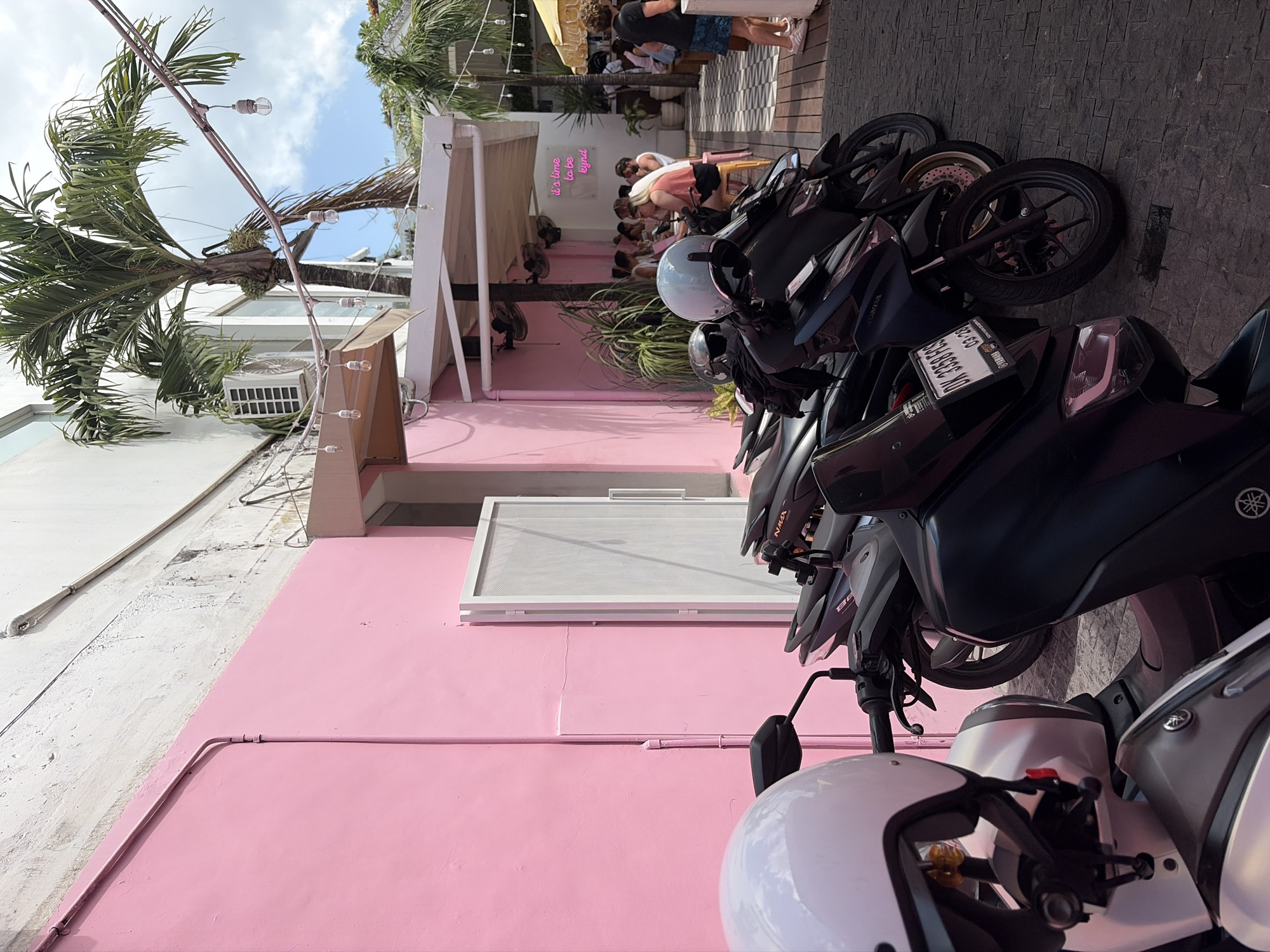 In North Kuta, Indonesia, a row of motorcycles is parked on a paved surface in front of a vibrant pink building. The building features white trim around its door and a large white door with a mesh inset. Above the building, palm trees and a cloudy sky are visible. String lights with exposed bulbs are strung across the scene. In the background, patrons are seated at tables in an outdoor seating area, with a neon sign on the wall reading "it's time to be kynd". A few people are standing and walking around the outdoor area.