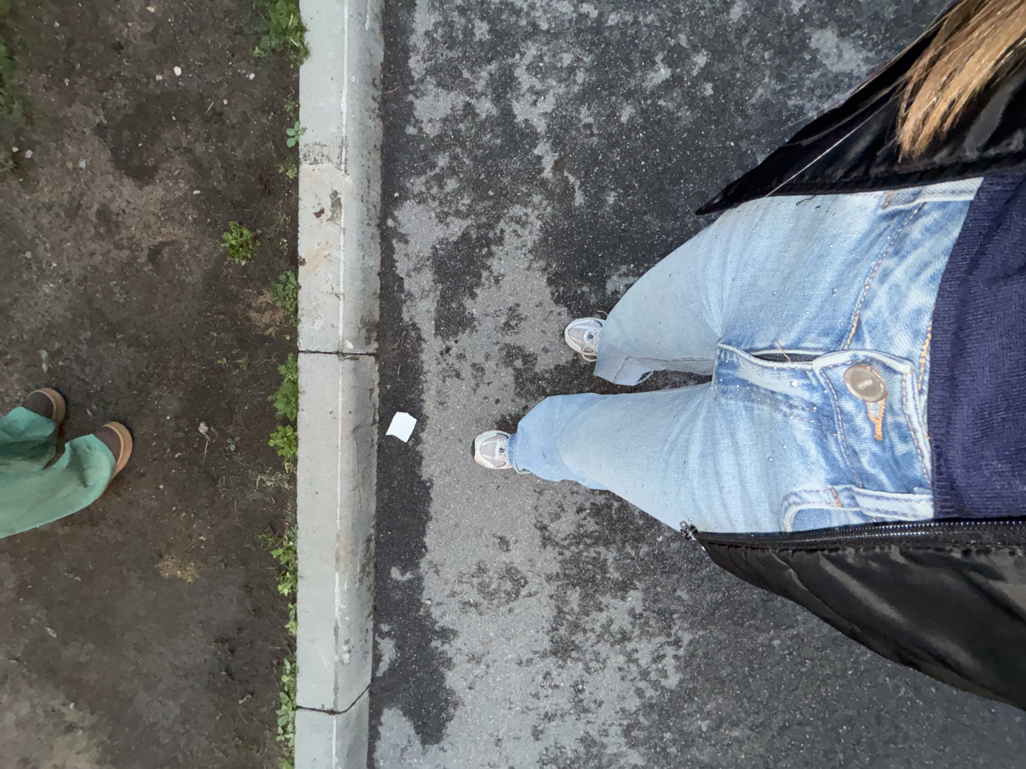 The image is a high-angle shot of a person wearing light blue jeans and white sneakers, standing on an asphalt surface with visible water spots. The person is also wearing a black jacket and a navy blue sweater. In the background, there is a concrete curb with some dirt and green vegetation on the other side. Further back, another person is visible, wearing green pants and brown shoes. The scene appears to be outdoors, possibly on a street or sidewalk. The location within Arfa, Russia is not specified in the image.