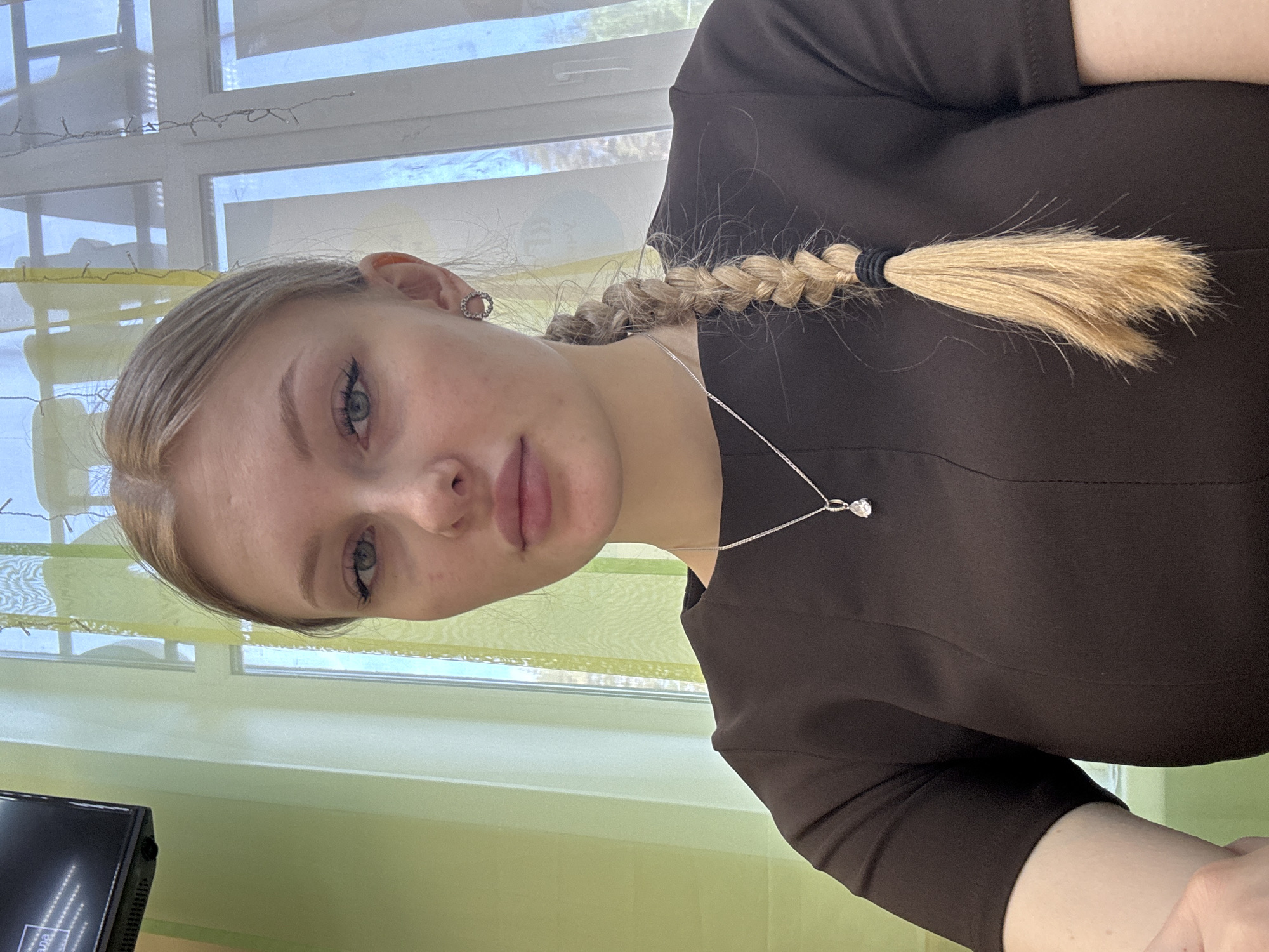 A young woman is depicted in a portrait. She has fair skin, blonde hair styled in a braid, and green eyes. She is wearing a dark brown garment and a silver necklace with a pendant. The background features a window with white frames and light-colored curtains. There is a monitor in the lower left corner. The location is likely indoors, in Perm, Russia, but the exact place cannot be determined from the image.