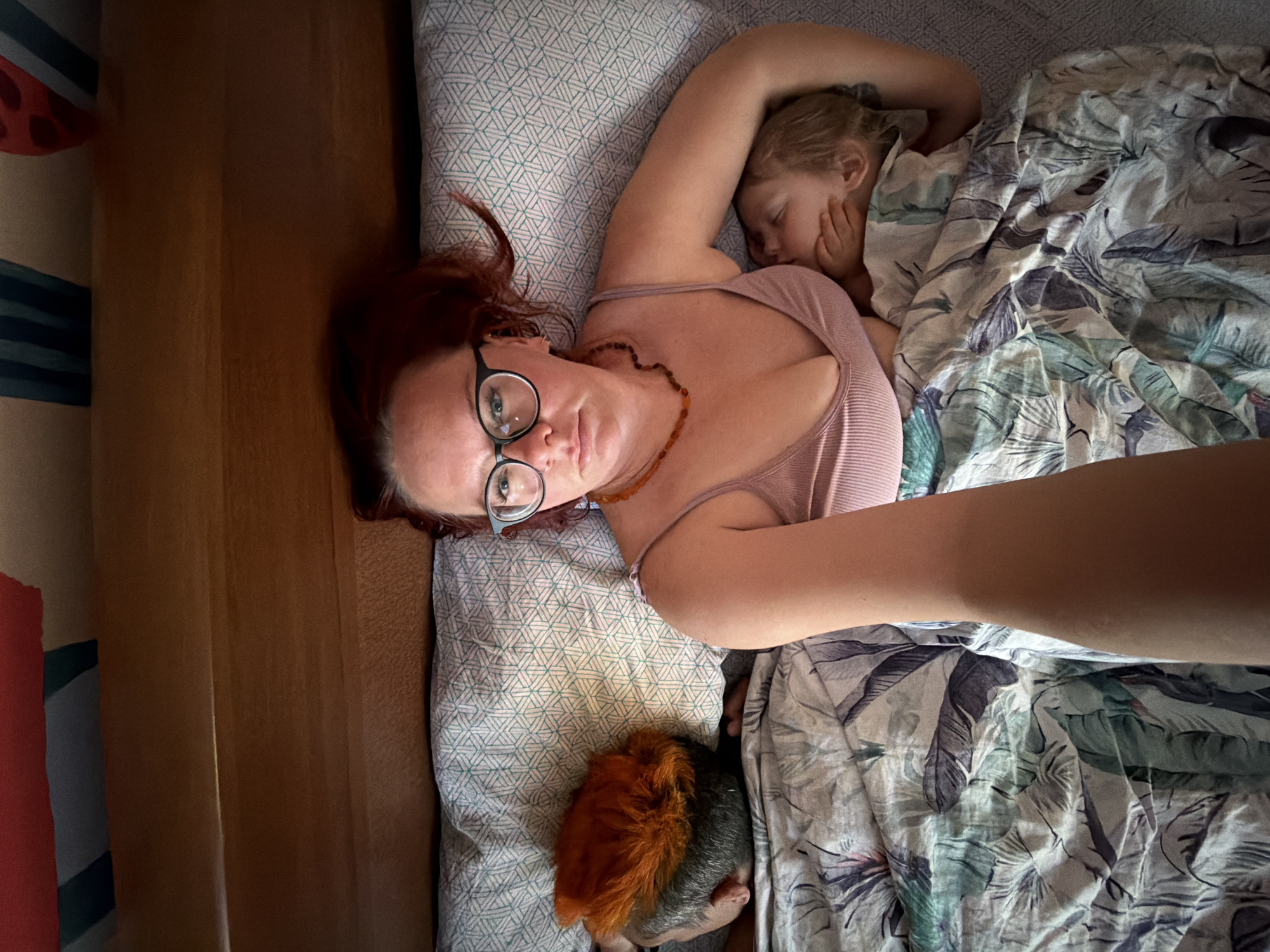 A woman with auburn hair and glasses lies on a bed in what appears to be a domestic setting. She is wearing a light pink tank top and a beaded necklace. Next to her, a young girl sleeps under a floral-patterned comforter. A pet rodent, possibly a guinea pig with brown and grey fur, rests on the pillow near the woman. The image appears to be a self-portrait taken from above, with the woman's arm extended. The location is unknown.