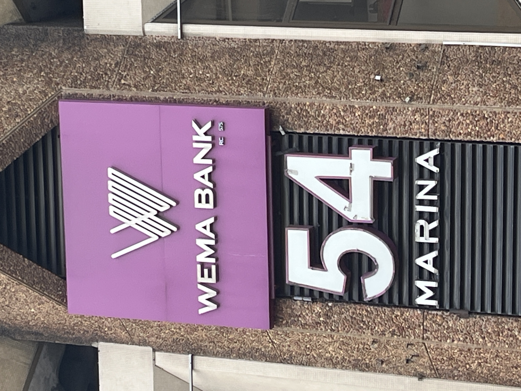 The image shows a WEMA Bank branch located at 54 Marina in Lagos, Nigeria. The bank's sign is purple with the bank's logo and name in white. Next to the bank sign is the address "54 Marina" in large white numbers and letters against a dark, vertically ribbed background. The building's facade is textured in a brown, pebble-like material. The image is taken from an angle, suggesting a position looking up at the building's signage.
