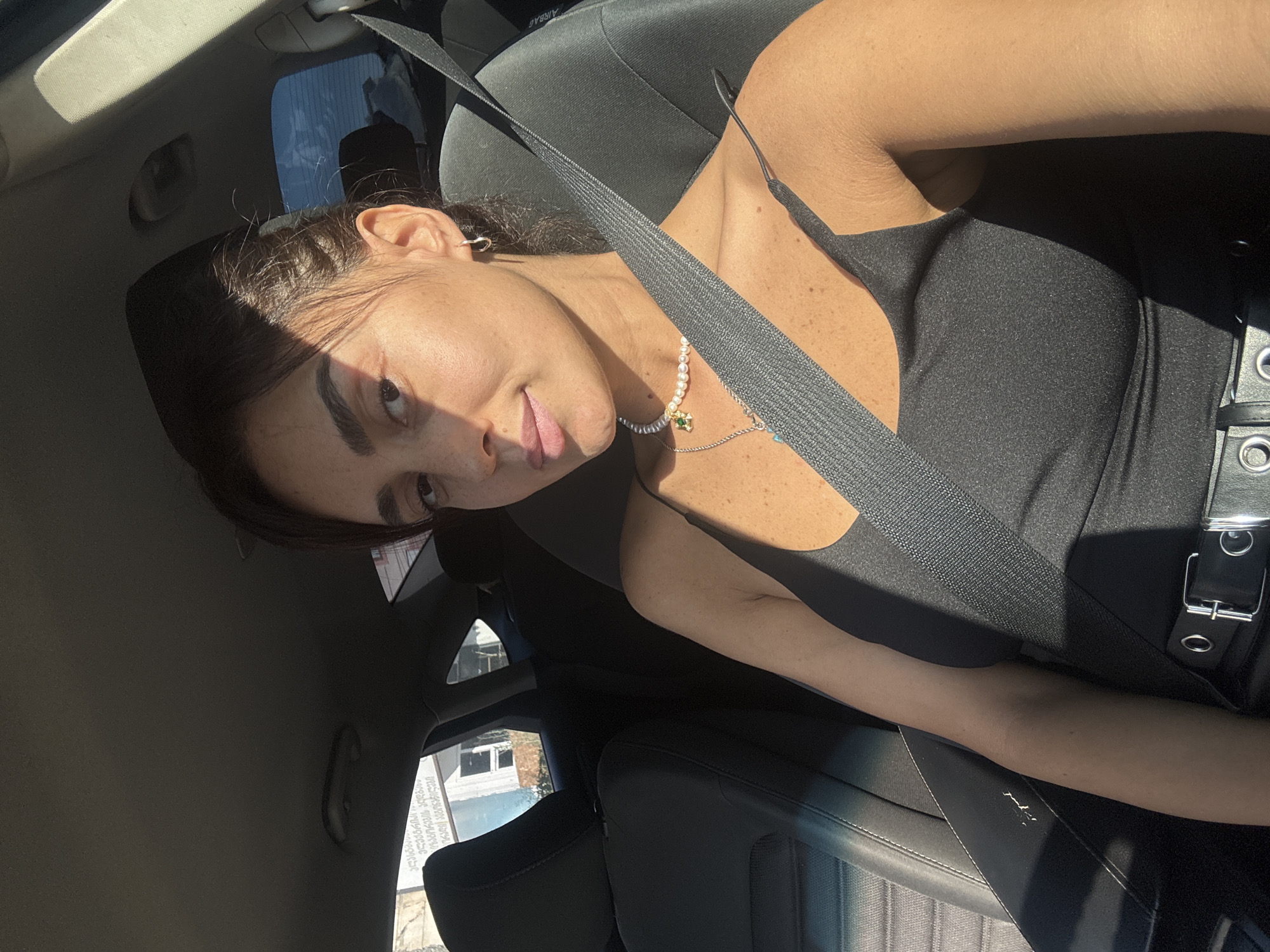 A young woman is sitting in the driver's seat of a car, her body angled towards the camera. She is wearing a black tank top, a pearl necklace, and a silver hoop earring in her left ear. A black seatbelt is fastened across her chest. The car appears to be parked, as indicated by the stillness of the scene. Sunlight streams in through the windows, casting shadows on her face and upper body. The background shows the interior of the car, including the dashboard and seats, and through the windows, a building with text in Georgian script is visible, suggesting the location is Tbilisi, Georgia.