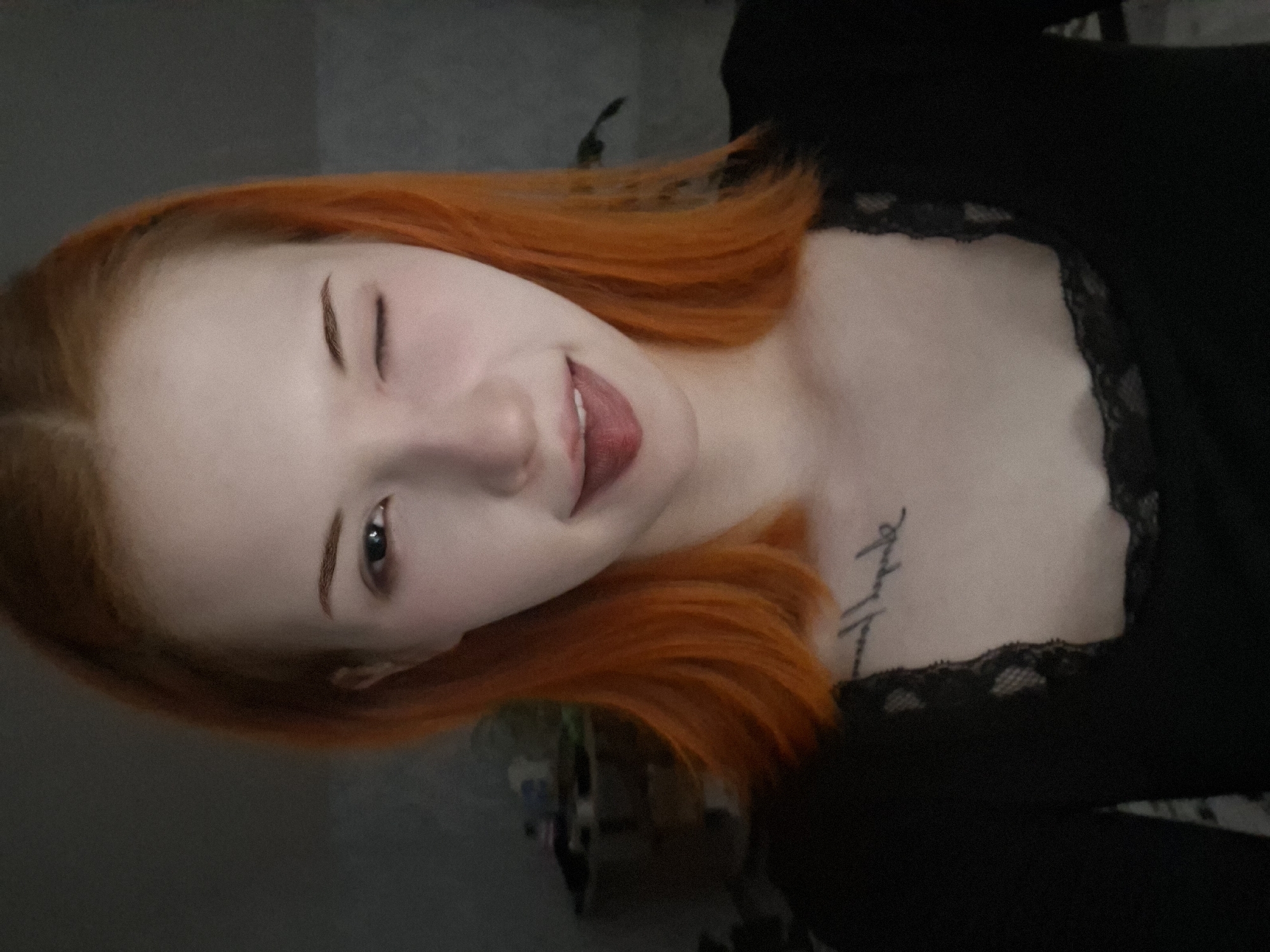 A young woman with red-orange hair is pictured indoors. She is wearing a black garment with lace trim along the neckline. Her right eye is closed in a wink, and she is sticking her tongue out slightly. A tattoo is visible on her chest. The location is likely somewhere in the city of Chaikovsky, Russia. The environment is dimly lit.