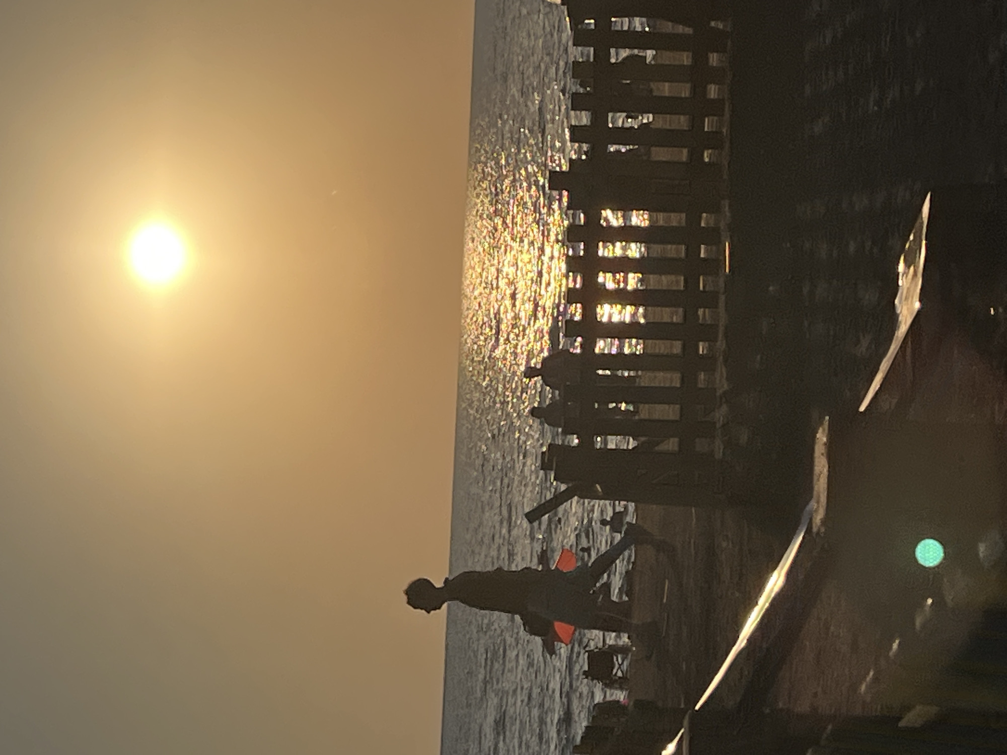 The image depicts a sunset scene at a beach in Ureki, Georgia. The sun is a bright, glowing orb low in the sky, casting a golden hue over the water and horizon. The sea is calm, with gentle waves reflecting the sunlight, creating a shimmering effect.
In the foreground, a wooden fence runs horizontally across the image. Silhouetted figures can be seen behind the fence, some appearing to be standing and others possibly sitting.
To the left of the frame, a person is walking along the beach, silhouetted against the bright sky. This individual is wearing shorts and a shirt, and carries a bright orange object under their arm. Further down the beach, other figures are visible, some wading in the shallow water. Beach chairs are also present on the sand.
The overall atmosphere is serene and peaceful, capturing the end of a day at the coast. The lighting suggests it is either sunrise or sunset.