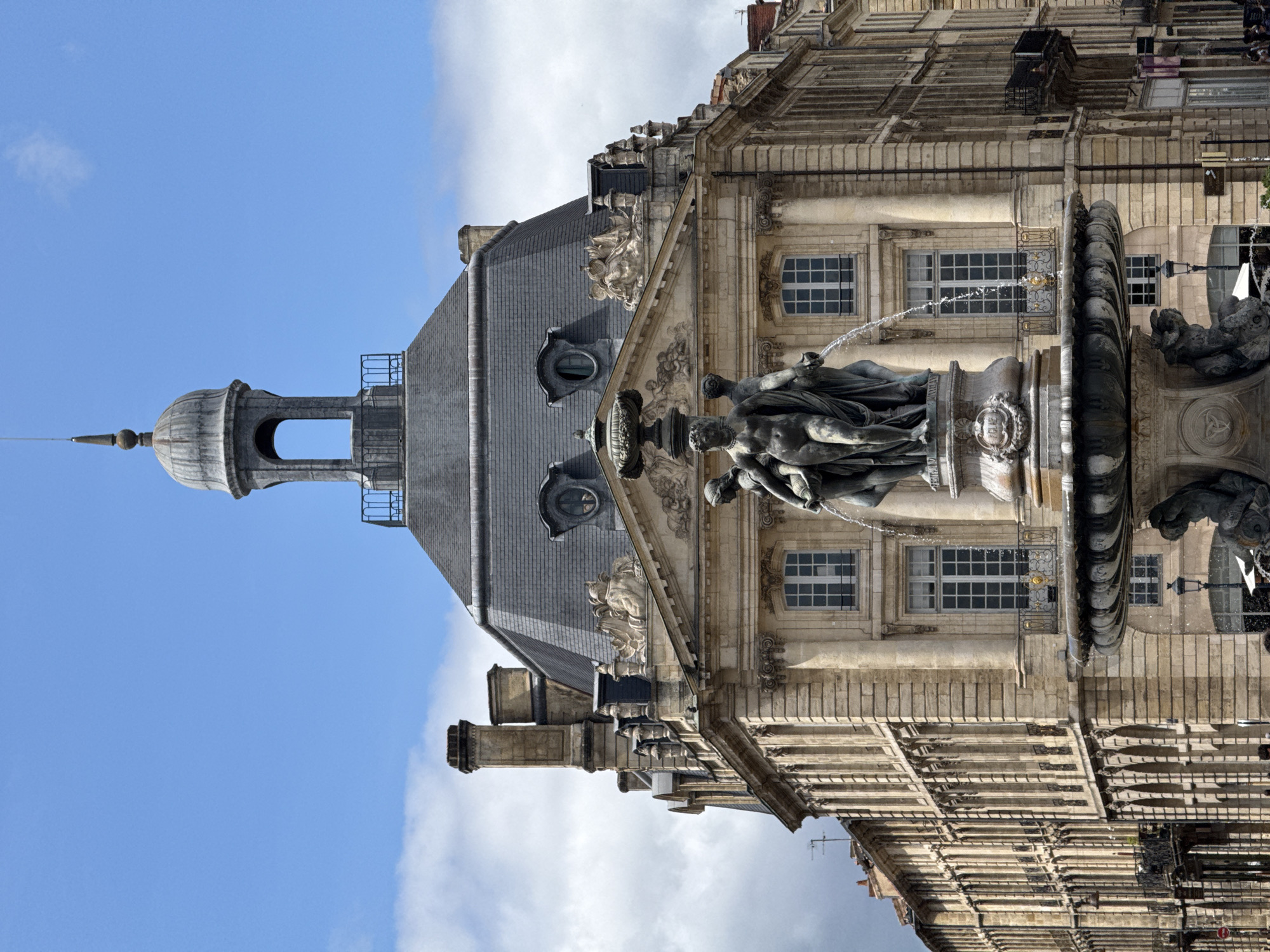 The image shows the Girondins Fountain, a prominent monument located in Bordeaux, France, specifically in the Place des Quinconces. The fountain features a multi-tiered structure with classical-style sculptures, including three female figures at the top pouring water from urns, and allegorical figures at the base. Behind the fountain, a grand stone building with a mansard roof and dormer windows dominates the scene. The architecture suggests a historical European city, consistent with Bordeaux's architectural heritage. The sky is partly cloudy, and the lighting indicates it is daytime. The image is captured from a slightly low angle, emphasizing the scale of the fountain and the building. There are no discernible people clearly visible in the foreground or mid-ground, though some blurry figures might be present at the base of the fountain or on the street in the distance. The overall scene depicts a public square with a significant historical monument and surrounding architecture.