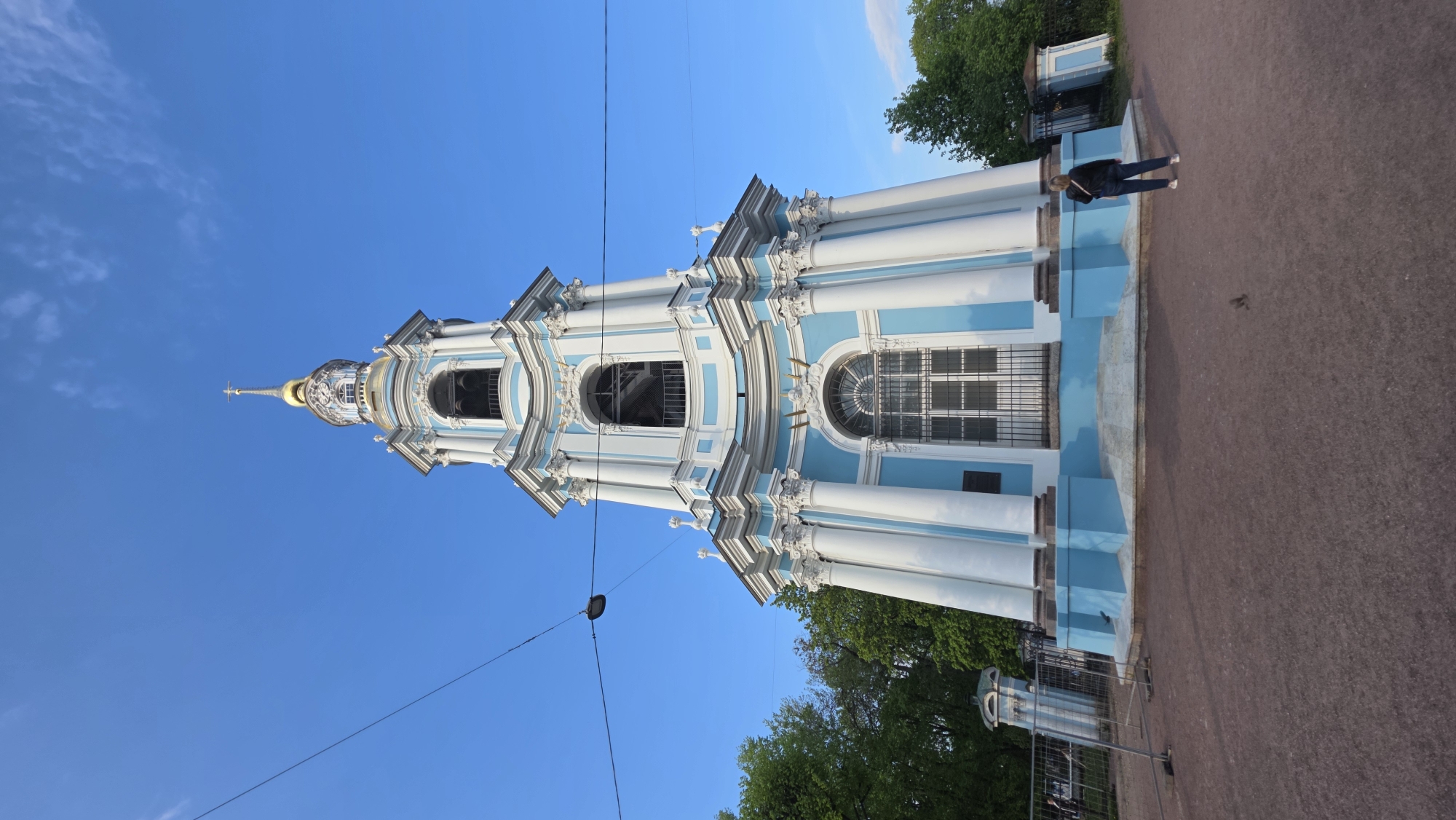 The image shows the Smolny Cathedral bell tower in Saint Petersburg, Russia. The bell tower is predominantly white with light blue accents. It features a golden dome and cross at its peak. White columns adorn its structure. The sky is a clear, bright blue. A person is seen walking near the base of the bell tower on a red brick path. Trees are visible at the base of the structure. Power lines run across the image.