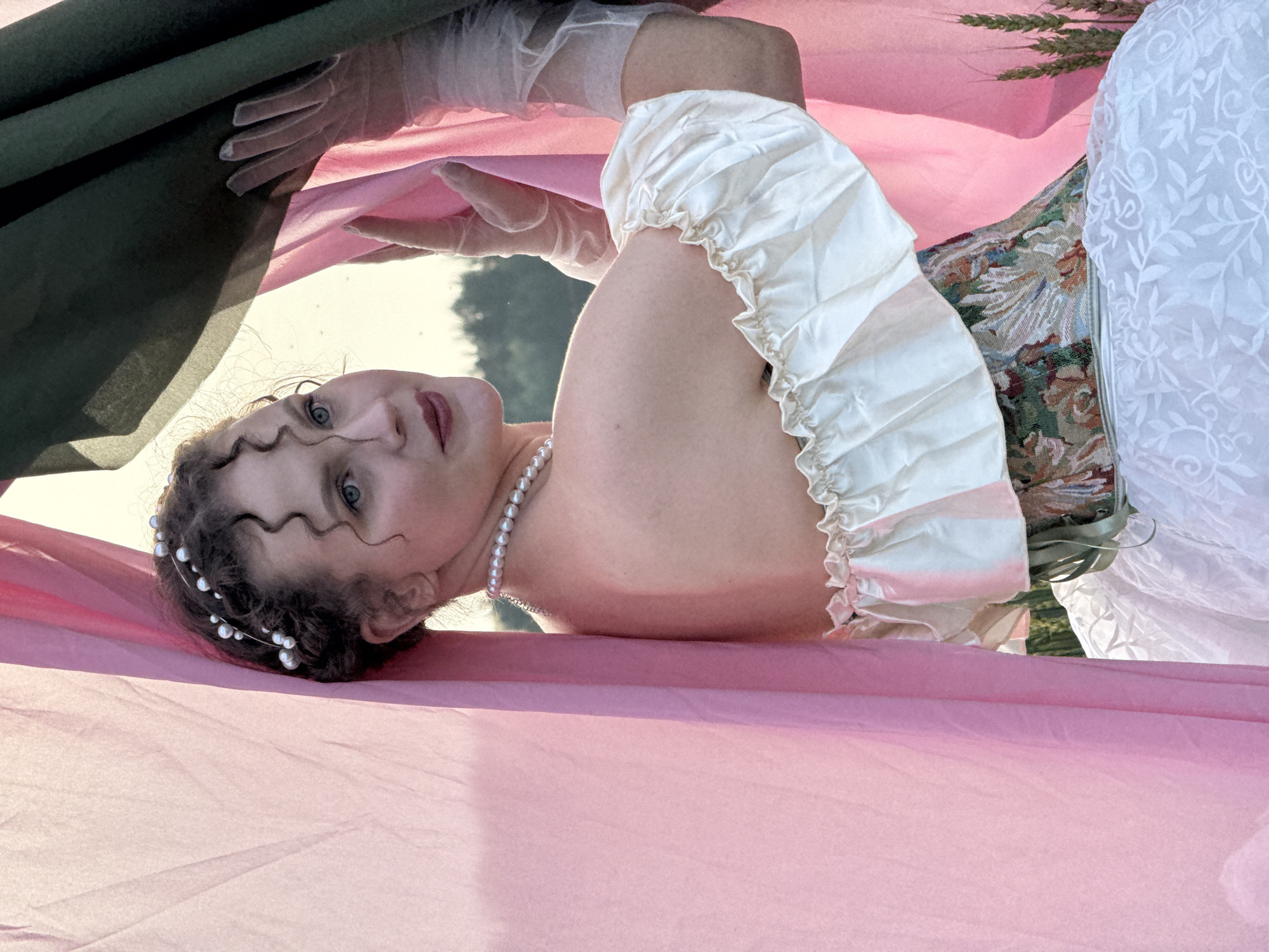 A person is lying down, covered by large pieces of fabric. The fabric is predominantly a soft pink, with a dark green fabric draped from above and a white, lacy fabric also visible. The person's head is tilted back, with their eyes open and looking towards the viewer. They have curly, dark hair adorned with a pearl headband. A pearl necklace is visible around their neck. The person is wearing a white, ruched garment with puffed sleeves. The scene appears to be outdoors, with hints of trees and a bright sky in the background, suggesting it is daytime. The lighting suggests it might be late afternoon or early evening. The overall mood is somewhat ethereal and dreamlike, with the fabrics creating a sense of enclosure. There is no visible text in the image. The location is Malyi Bukor, Russia.