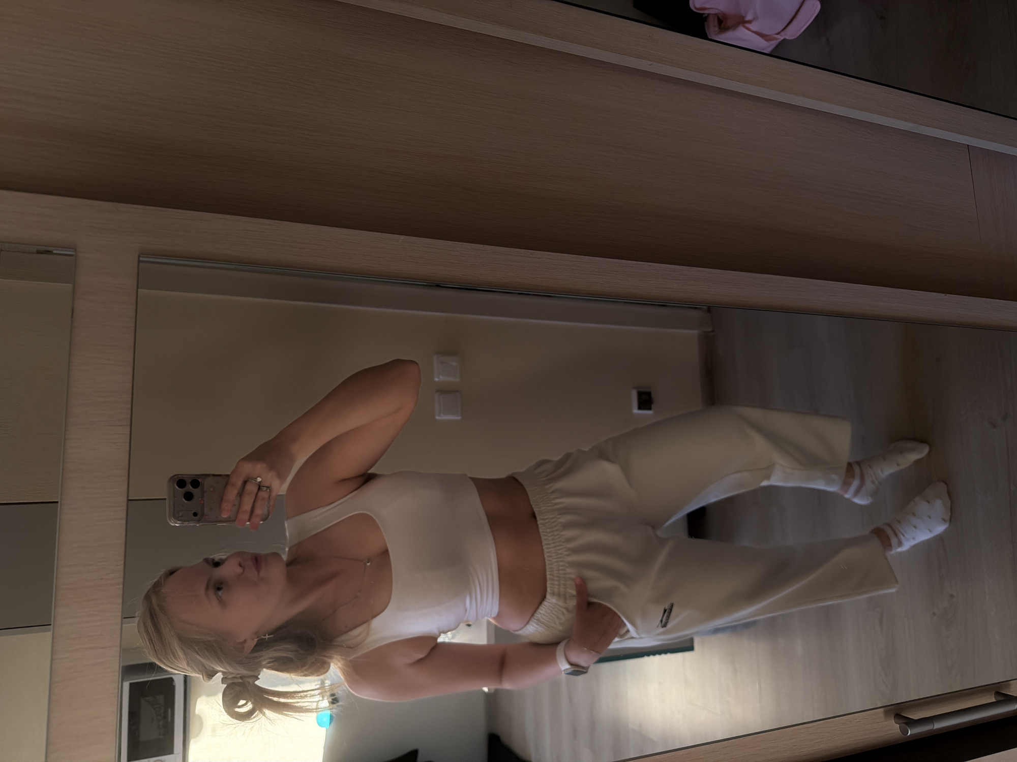 The image displays a young woman taking a full-length mirror selfie in an indoor setting, likely a home. She has blonde hair styled in a ponytail and is looking at her smartphone, which she holds in her left hand. The phone has a patterned case and visible multiple camera lenses. She is wearing a white sleeveless crop top, light-colored (possibly off-white or beige) baggy sweatpants, and white socks with subtle striped or dotted patterns. On her left wrist, she wears a white smartwatch, and a ring is visible on her left hand. A thin necklace with a small pendant, possibly a cross, adorns her neck.
The background, largely reflected in the mirror, reveals a modern interior. Light-colored wooden cabinets or wardrobe doors are visible above and to the sides of the mirror. The floor is made of light-toned wood or laminate. Reflected in the mirror, one can see white wall-mounted electrical outlets or switches and a microwave or a similar small appliance. Above the mirror, a pink item, possibly folded clothing, is perched on a shelf. Below the mirror, two silver handles suggest a drawer or cabinet.
The woman is posing casually, with her right hand resting on the waistband of her sweatpants. The lighting suggests a daytime setting. No clear text is legible on her phone case or on the small label on her sweatpants. The scene captures a personal moment in a contemporary domestic environment.