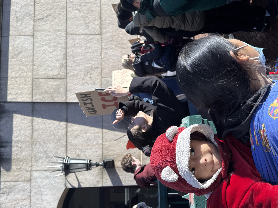 A protest scene captured from a high angle. A child in a red hooded jacket looks up at the camera. A woman wearing a mask stands close by. Behind them, a person holds a sign that reads "Abolish ICE". Other protesters are visible in the background, some wearing winter clothing. A decorative lamp post stands near the front. The ground appears to be made of stone or tile.