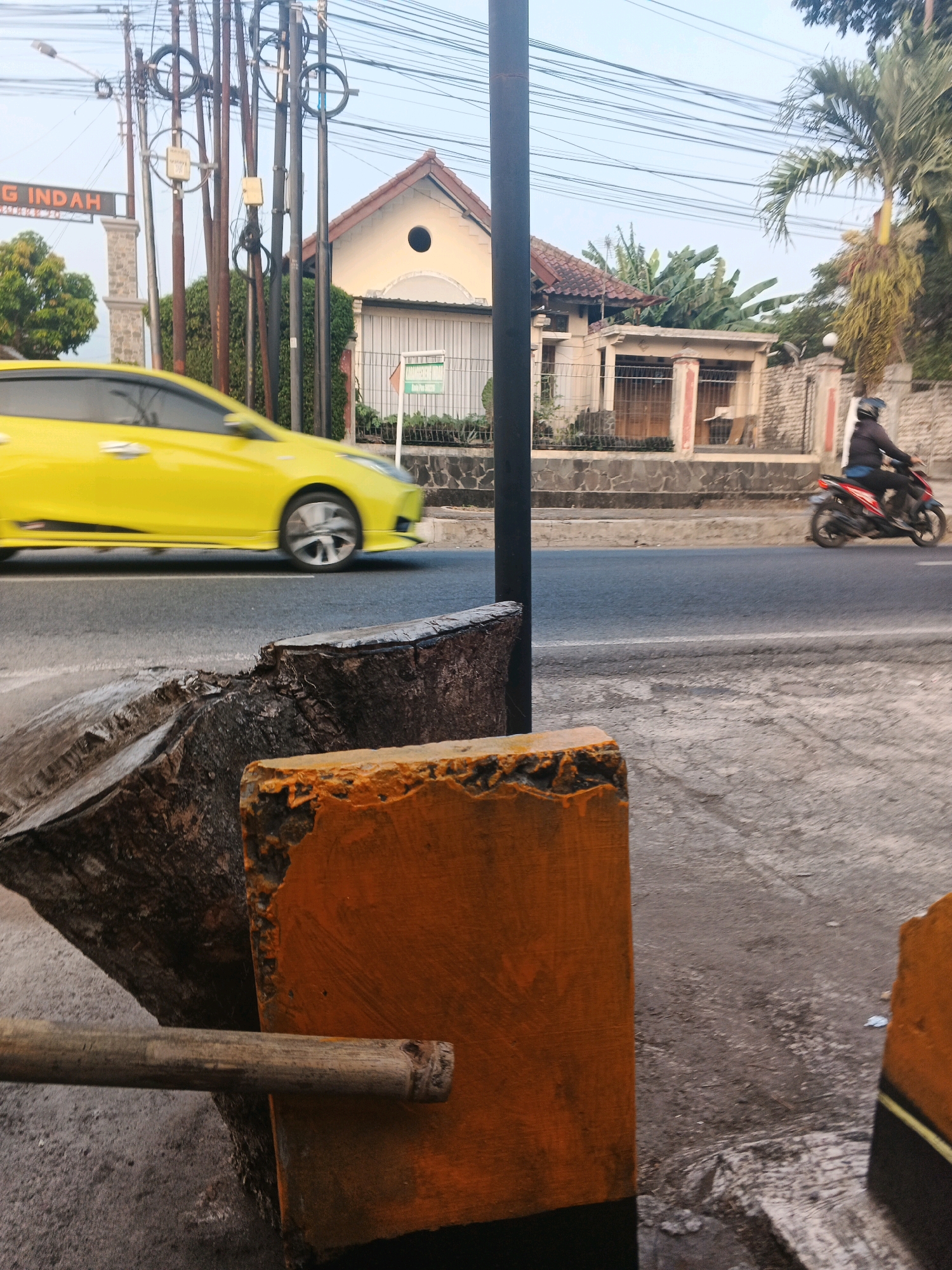 The image captures a street scene, likely in Semarang, Indonesia, characterized by a mix of urban infrastructure and elements of everyday life. In the foreground, the image is dominated by rough-hewn logs and brightly painted orange structures, possibly part of a roadside barrier or temporary construction. A bamboo pole rests horizontally across these elements. Beyond this, a road runs horizontally across the frame, with a bright yellow car in motion, its wheels blurred, indicating speed. Further down the road, a person is riding a red and black motorcycle, also in motion. In the background, a line of utility poles with numerous wires crisscrosses the sky, typical of Indonesian urban settings. Behind the poles, a house with a red-tiled roof and a distinctive arched window is visible, surrounded by lush green trees, including banana trees and a palm tree. The overall lighting suggests it is either early morning or late afternoon, with a clear sky. The environment appears to be a moderately busy street, with evidence of both residential and public spaces.