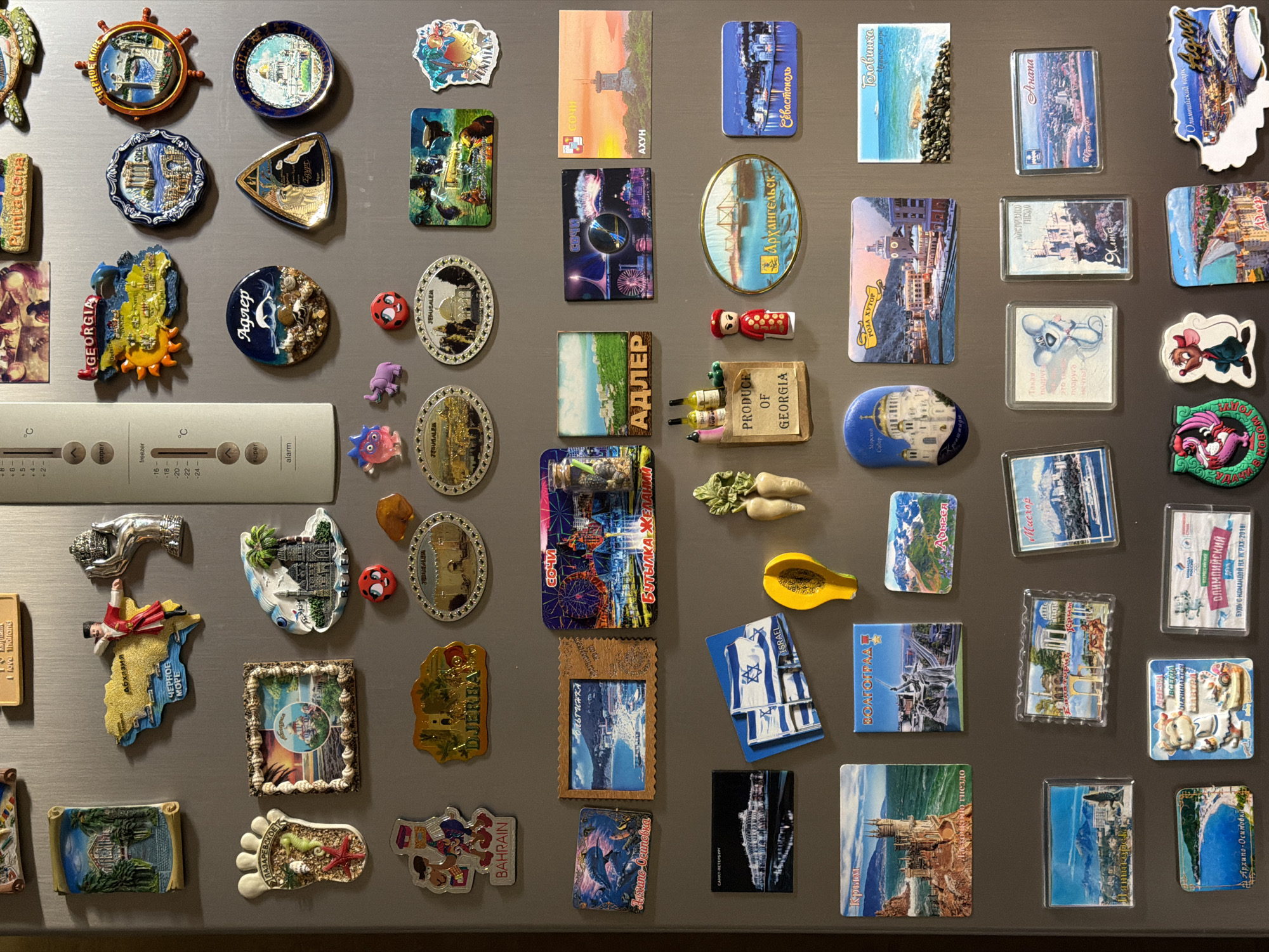 This image captures a close-up, overhead view of a refrigerator door covered with a diverse collection of magnets. The metallic grey surface of the refrigerator is visible, along with its digital control panel.
The main subjects are the numerous souvenir and novelty magnets, varying widely in size, shape (rectangular, oval, circular, irregular 3D), and material (plastic, metal, ceramic, resin). They are arranged in a dense, almost mosaic-like pattern.
Many magnets are travel souvenirs, depicting landmarks, cityscapes, natural scenery, and cultural symbols from various locations. Visible locations and themes include: GEORGIA (multiple times), Agrep, Jerusalem, Адлер (Adler), Сочи (Sochi), АХУН (Akhun), Архангельск (Arkhangelsk), Севастополь (Sevastopol), Головинка (Golovinka), Анапа (Anapa), Мисхор (Miskhor), Ялта (Yalta), РОЗА ХУТОР (Roza Khutor), КРЫМ (Crimea), САНКТ-ПЕТЕРБУРГ (Saint Petersburg), ВОЛГОГРАД (Volgograd), ISRAEL, DJERBA, BAHRAIN, Архипо-Осиповка (Arkhipo-Osipovka), Punta Cepa, Egypt, love Thailand, Лазаревское (Lazarevskoye), Адыгея (Adygea), and "ЧЕРНОЕ МОРЕ" (Black Sea). Specific items shown include churches, castles, lighthouses, beaches, traditional figures, and local produce like wine and roots.
Novelty magnets feature various characters and objects, such as a turtle, a fish, a ladybug, purple creatures, a cartoon mouse, a human-like figure, a ship's wheel, a foot with seashells, and a yellow fruit. One magnet reads "БУТЫЛКА ЖЕЛАНИЙ" (Bottle of Wishes). Another, related to the 2014 Sochi Olympics, states "ОЛИМПИЙСКИЙ БУДЬ С КОМАНДОЙ НА ИГРАХ-2014" (Olympic Be with the team at the 2014 Games). A cow-shaped magnet has text "ВСЕГДА НАЧИНАЕТСЯ" (Always begins).
At the bottom left, a refrigerator control panel is visible, displaying temperature settings: "+5", "+4", "+2", "°C" for the fridge, and "-16", "-18", "-20", "-22", "-24", "°C" for the freezer. Buttons for "super freezer" and "super alarm" are also present. The lighting is bright and even, highlighting the details and colors of the magnets.