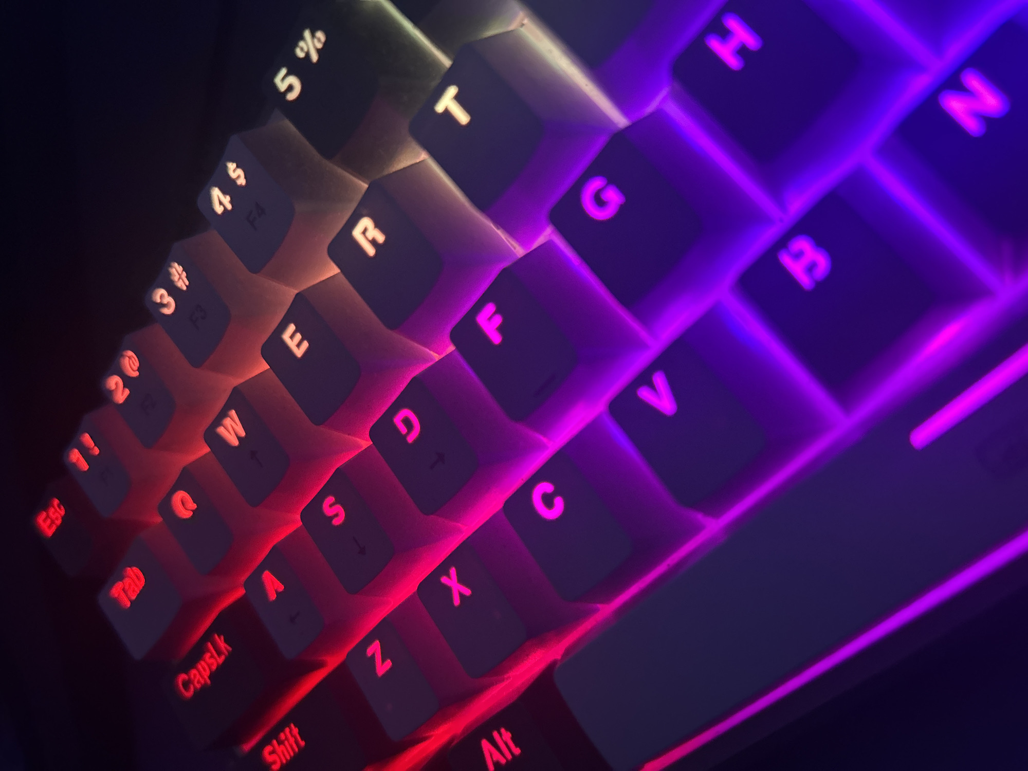 The image is a close-up, high-angle shot of a backlit keyboard with a gradient of red, purple, and white lighting. The keys are dark grey with glowing white or red lettering. Visible keys include "Esc", "Tab", "CapsLk", "Shift", "Z", "X", "C", "V", "F", "G", "B", "R", "T", "H", "N", "1!", "2@", "3#", "4$", "5%". The lighting creates a dramatic and futuristic effect. There are no people, animals, or signs of a city visible in the image. The image appears to be a still life of a computer peripheral.