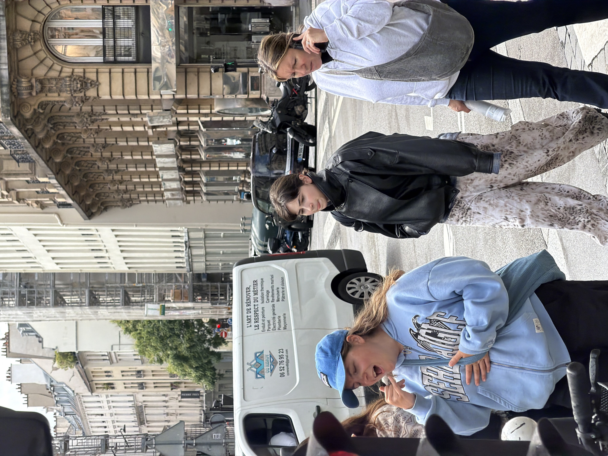 The image shows three people on a city street with buildings in the background. A woman with dark hair in a black leather jacket stands in the middle. Another woman with blonde hair is to the right, wearing a white sweatshirt and dark pants and holding a water bottle. To the left, a young woman wearing a blue hooded sweatshirt and a blue baseball cap appears to be eating. A white van with French text and a phone number on the side is visible. The buildings in the background suggest the image was taken in Paris, France.