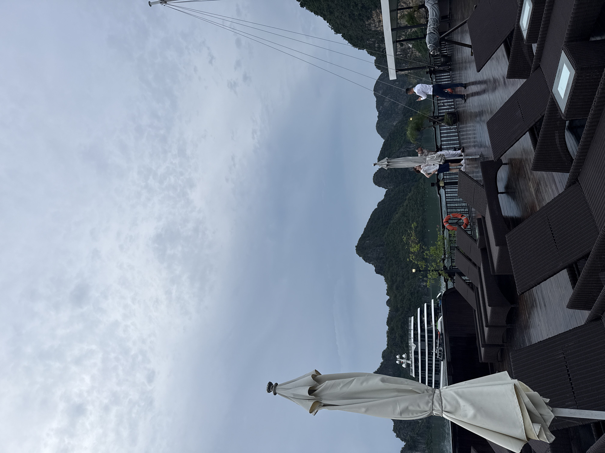 The image shows a view from the deck of a boat or cruise ship in Ha Long Bay, Vietnam. The scene is set against a backdrop of lush green karsts rising from the water, characteristic of the location. The sky is overcast with scattered clouds, suggesting a potentially damp or moody atmosphere.
In the foreground, a folded white umbrella stands prominently, tied with a rope. The deck is made of dark wood and is wet, indicating recent rain or mist. Several dark brown wicker lounge chairs and tables are arranged on the deck, some with lit white square lamps on them, providing a touch of ambient light.
In the middle ground, two people are visible. One, wearing a white shirt and dark pants, is standing near the railing, looking out at the scenery. Another individual, dressed in white, is also standing near the railing, engaged in conversation with someone out of frame. Further down the deck, a group of people appear to be gathered, though their details are less clear. A life buoy is visible attached to the railing.
In the background, a modern, multi-decked cruise ship with a distinctive white superstructure and black-tinted windows is moored or sailing in the bay. The karsts are heavily forested and appear to be shrouded in mist, adding to the dramatic and natural beauty of the setting. The overall mood is serene, with a hint of tranquility despite the overcast weather. No visible text is present in the image.