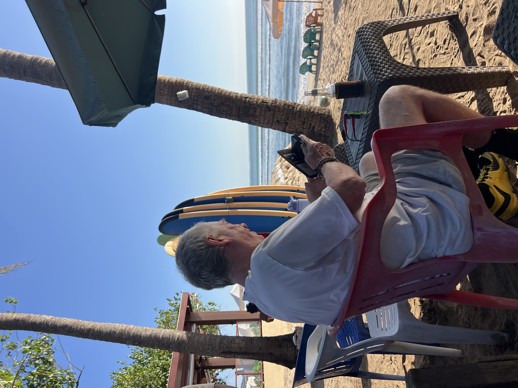 An elderly Caucasian man with graying hair sits in a red plastic chair on a sandy beach in Kuta, Indonesia, facing away from the camera. He is wearing a white t-shirt and gray shorts and is looking down at a tablet device he holds in his hands. A bottle of dark liquid and a pair of red-framed glasses rest on a woven plastic table in front of him. Behind the man, two surfboards are propped up against a building. The ocean with gentle waves is visible in the background, with several empty beach chairs on the sand. A large palm tree trunk stands to the right of the man, with a green umbrella extending from behind it. The sky is clear and blue.
