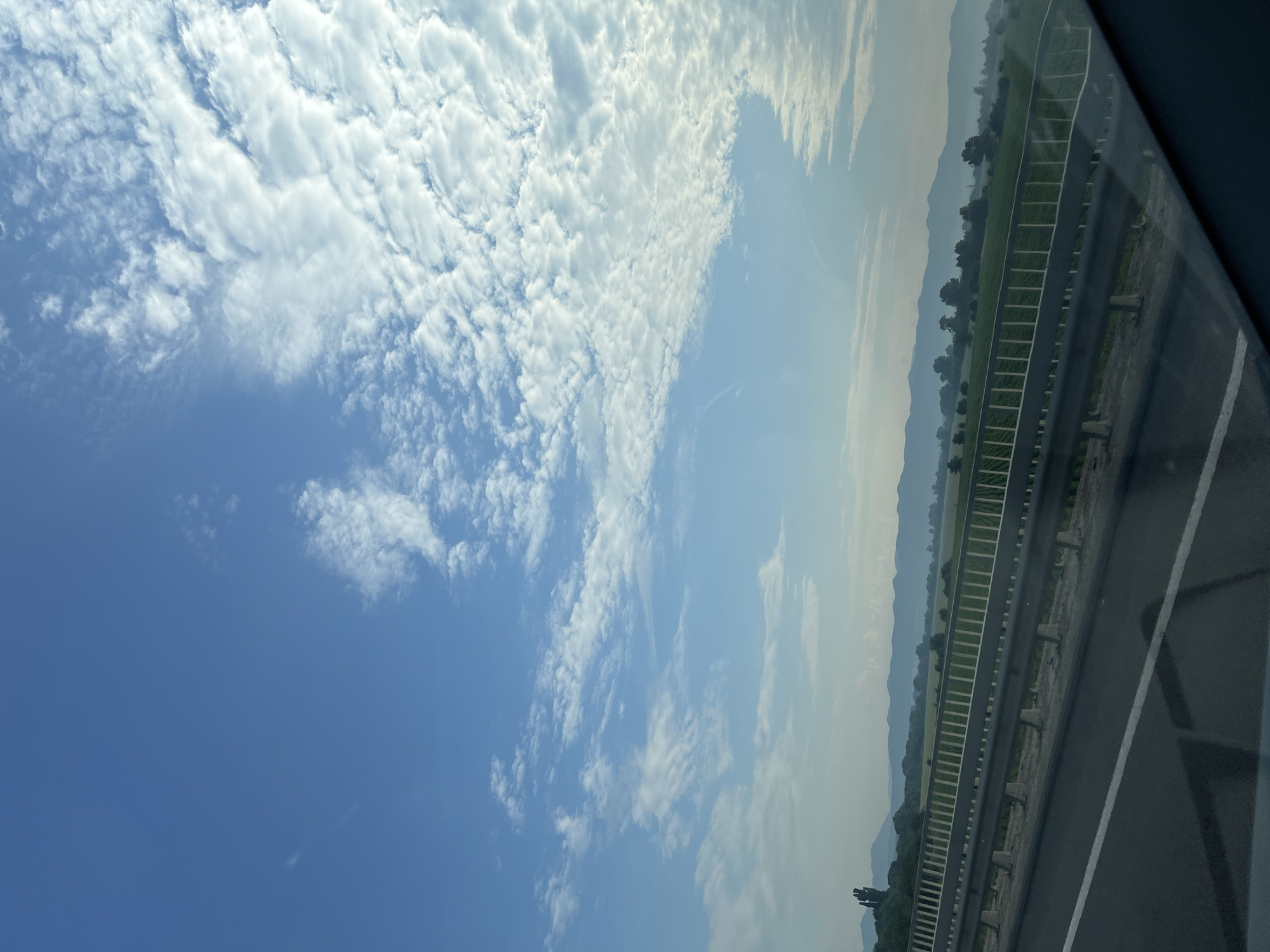 The image depicts a scenic view from a highway, looking out over a rural landscape. The sky is a vibrant blue, filled with a mix of fluffy white clouds and wispy cirrus formations. In the foreground, a silver guardrail runs along the side of the road, with a dark asphalt roadbed visible below. Beyond the guardrail, a green field stretches out, dotted with trees and culminating in a distant range of rolling, hazy mountains. The overall impression is one of tranquility and expansive natural beauty.