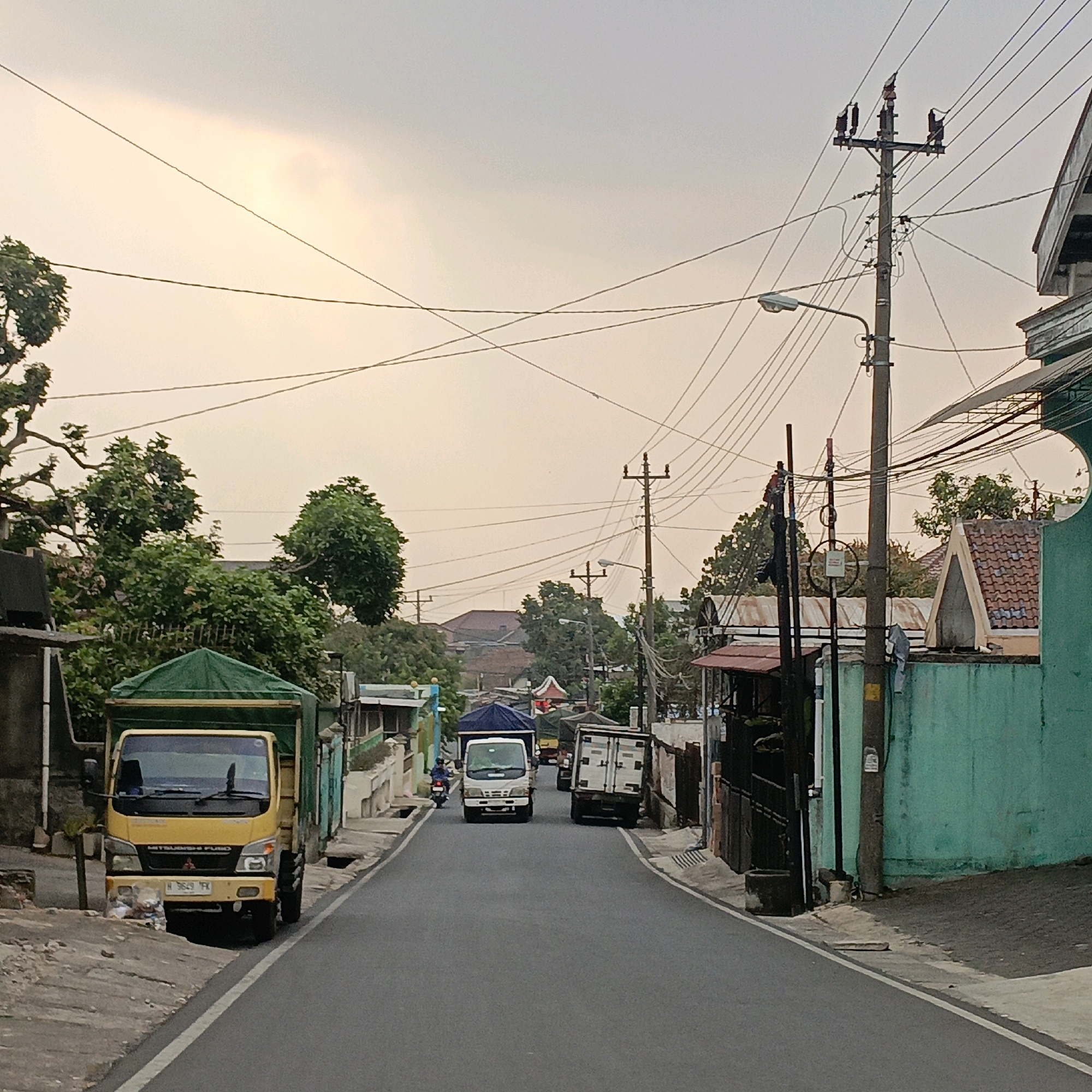 The image shows a street scene in Salatiga, Indonesia, under an overcast sky. In the foreground, a yellow Mitsubishi Fuso truck is parked on the left side of the road, with its headlights and license plate visible. The road is paved and has a white line marking its edge. Further down the street, two white trucks are driving away from the camera, with a motorcycle passing them on the left. The street is lined with buildings, some of which appear to be houses with tiled roofs, and others are commercial establishments with metal gates. Green trees and foliage are present throughout the scene, adding to the urban landscape. Electrical poles with numerous wires crisscross the sky, indicating a developed infrastructure. The overall impression is a typical street scene in a bustling Indonesian city.