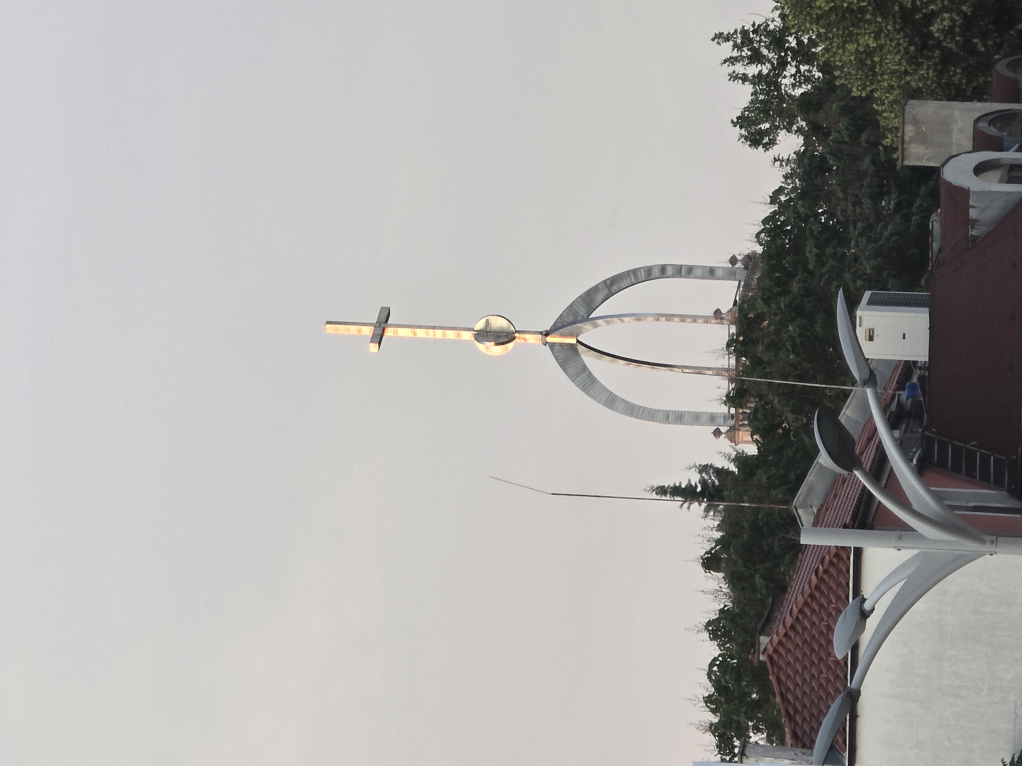 The image displays a prominent modern metallic sculpture, likely photographed during dawn or dusk due to the soft, muted light of the sky. The central feature is a stylized cross, seemingly illuminated or reflecting light, positioned at the top of a large, abstract structure composed of several wide, curving metal arcs that resemble a crown or a gateway. A spherical element connects the cross to this base.
Behind the sculpture, dense green foliage from trees is visible, partially obscuring parts of buildings with distinctive reddish-brown tiled roofs. White objects, possibly air conditioning units or satellite dishes, are discernible on these rooftops. In the foreground on the right, several contemporary street light poles with multiple curved arms and light fixtures ascend into the frame.
The sky is clear and bright without clouds, contributing to the serene atmosphere. No people or specific activities are depicted, making it a static scene focused on the monument and its immediate surroundings. There is no visible text. The setting is an urban environment, indicative of a landmark in a city such as Plovdiv, Bulgaria, where a similar Monument of the Holy Cross exists.