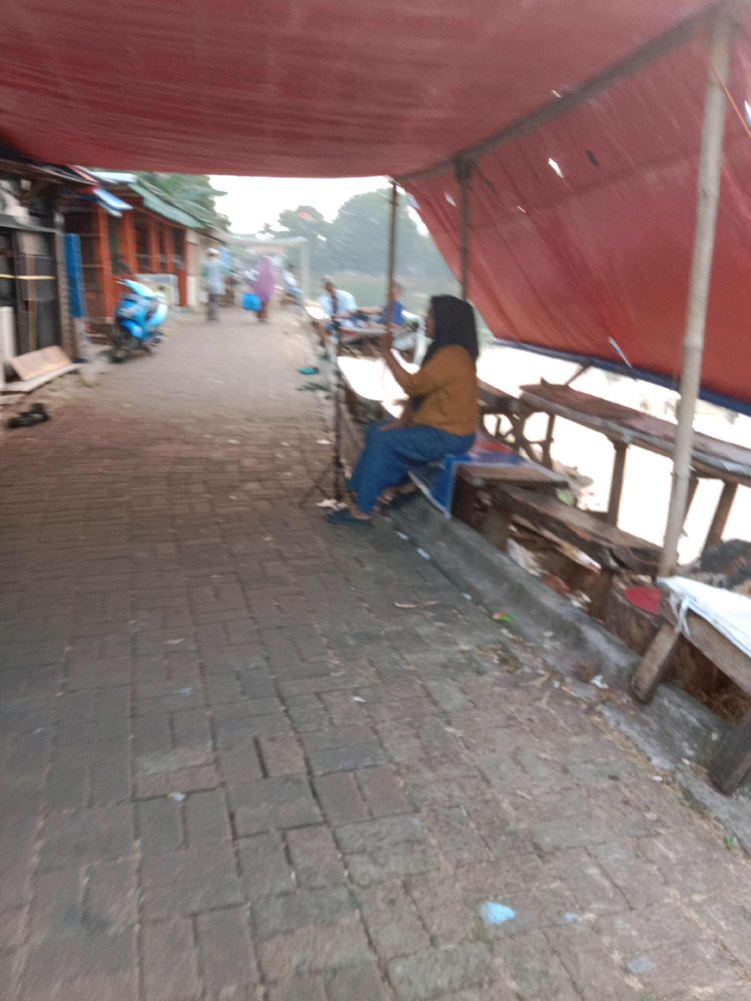 The image depicts an outdoor scene under a red tarp canopy, possibly a market or roadside stall area. A woman wearing a mustard-colored shirt and blue pants sits on a bench. She is looking to her right, and her right hand is raised, holding a small, dark object, possibly a phone. In the background, several people are walking along a paved pathway. A blue motorcycle is parked to the left, and there are wooden structures and tables visible along the right side of the pathway. The ground is paved with rectangular bricks. The overall scene suggests a warm, possibly humid environment with diffused natural light. The location is identified as South Tangerang, Indonesia.