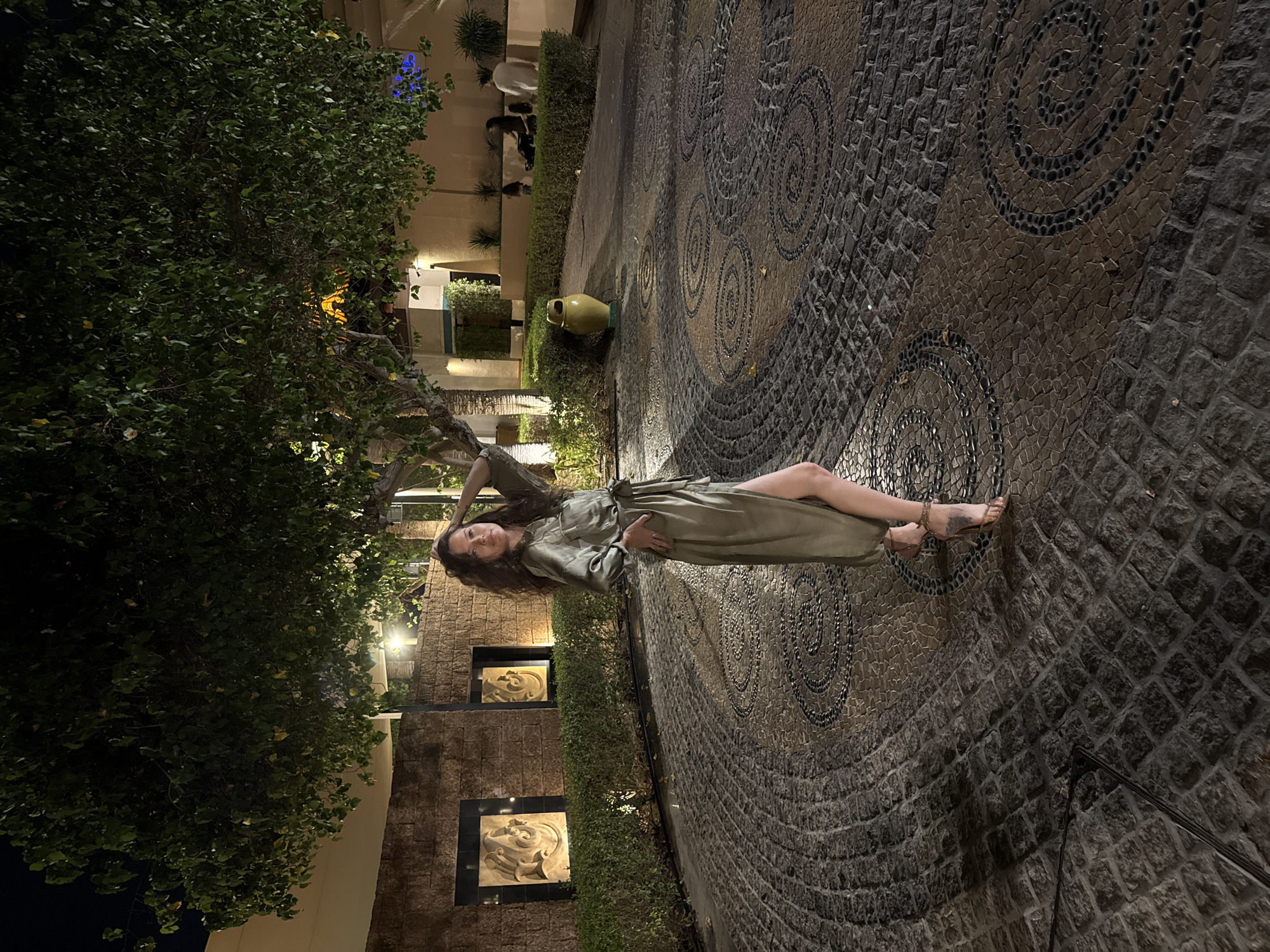 This image, captured in Dubai, United Arab Emirates, depicts an outdoor night scene featuring a single female subject in a landscaped courtyard.
The primary subject, an adult female with long, wavy dark brown hair, stands slightly left of center, angled towards the right side of the frame, with her face turned towards the camera. She wears a long-sleeved, olive-green collared maxi dress with a tie-waist and a high left-side slit. Her left leg is partially exposed, and she wears light-colored open-toed high-heeled sandals with ankle straps. Her left hand rests on her hip, while her right hand is raised near her head.
The background on the left features a dark-colored, textured stone wall with two rectangular recesses, each containing an illuminated light-colored sculptural relief. A manicured green hedge runs along the base of this wall and continues towards the right, forming a border for a path. To the right of the hedge, a yellow urn-shaped object is visible. Further back, on the right, additional individuals are dimly visible, including at least two adults and possibly a child or a stroller, appearing to be in an outdoor seating area. Architectural structures with warm and cool artificial lighting (including a blue light source) are present in the distant background. A large, densely leafed tree occupies the upper right portion of the frame, with visible illumination on its leaves.
The ground is composed of an intricate mosaic pavement. Dark grey cobblestones form segments, interspersed with lighter brown/tan sections made of smaller, irregularly shaped stones. Within these lighter sections, distinct spiral patterns are created using small, dark pebbles. A dark, narrow metal strip or cable extends diagonally across the bottom left corner of the frame. The scene is illuminated by multiple artificial light sources, indicating nighttime.