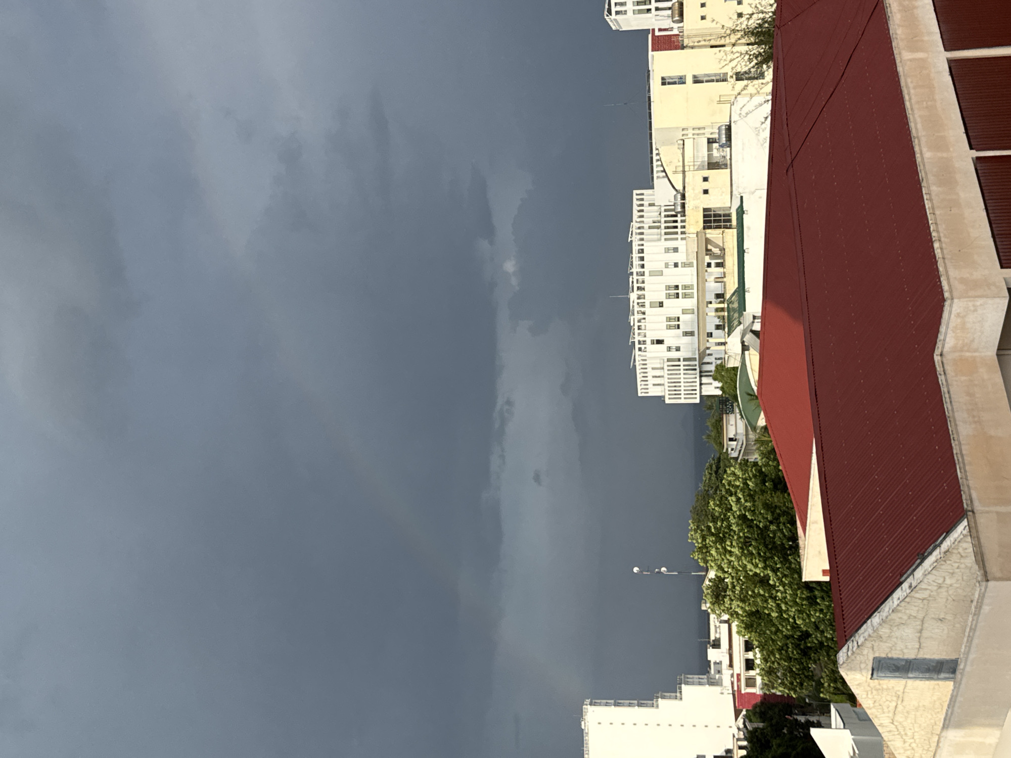 The image shows a city skyline with a dark, cloudy sky. A faint rainbow is visible in the upper left portion of the sky. Several buildings are in the foreground and midground, including a modern white building with many windows and a building with a red roof. Green trees are also visible. The image appears to be taken from a high vantage point, possibly a building overlooking the city. The overall impression is one of an impending storm or the aftermath of rain.