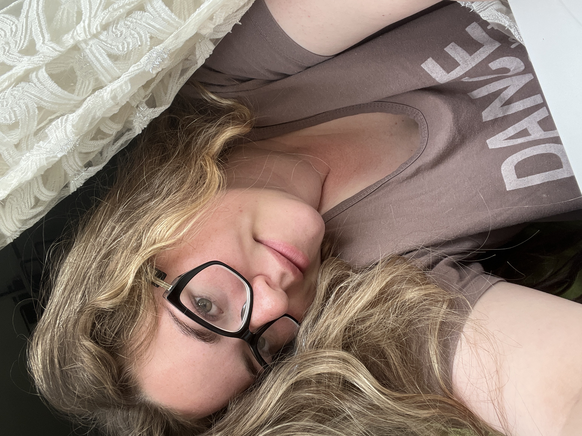 A fair-skinned woman with long, light-brown curly hair and glasses is lying down. She is wearing a brown t-shirt with the word "DANCE" printed on it. A white lace blanket is partially covering her. The location is not specified.