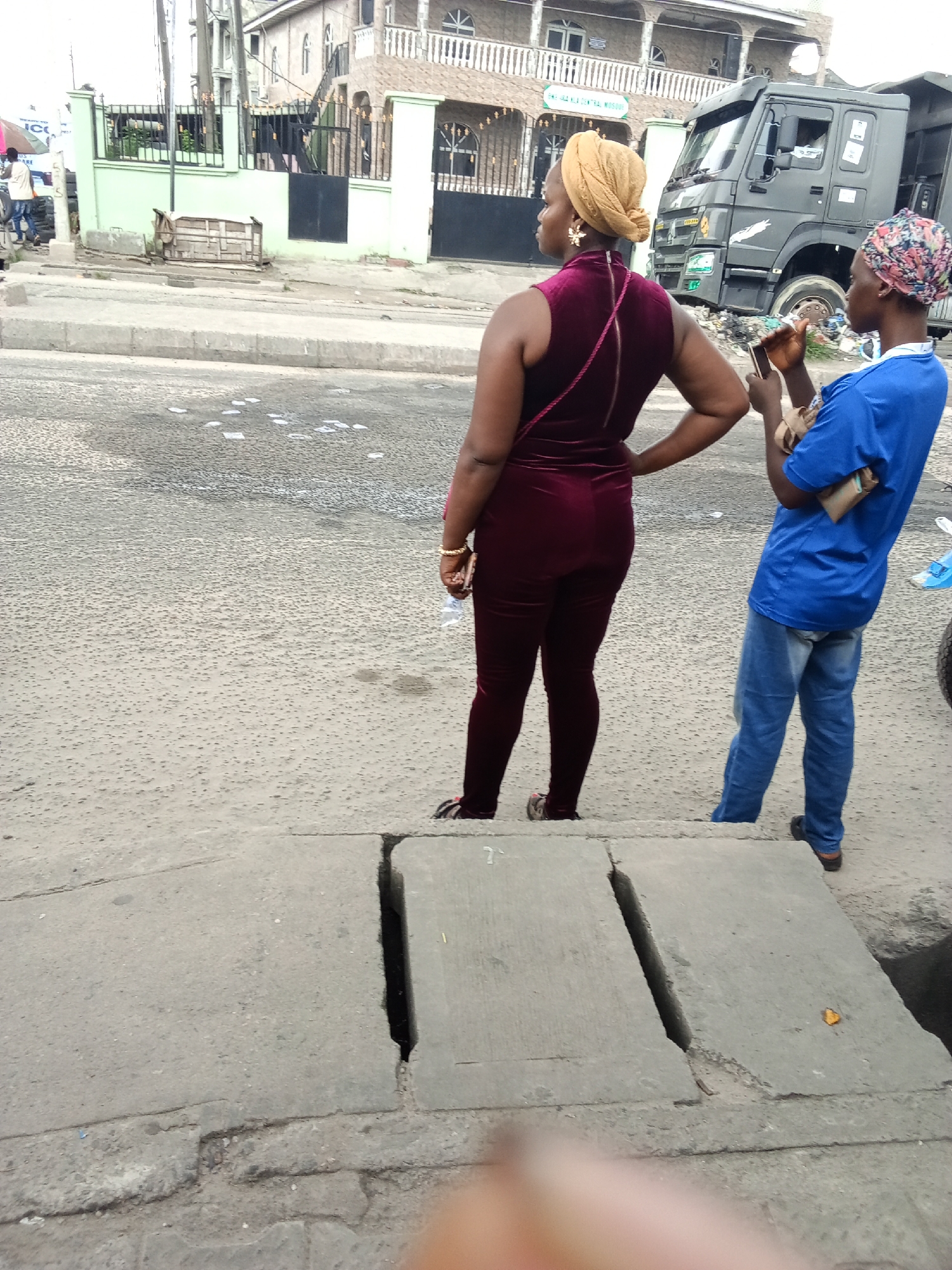 Two women stand on a concrete curb beside a street in Ado, Nigeria. The woman on the left is wearing a maroon jumpsuit and a tan headwrap. She is looking to the left with her hand on her hip. The woman on the right is wearing a blue shirt, jeans, and a patterned headwrap. She is holding a cell phone to her ear. Behind them is a large truck and a building. Debris is visible on the street.