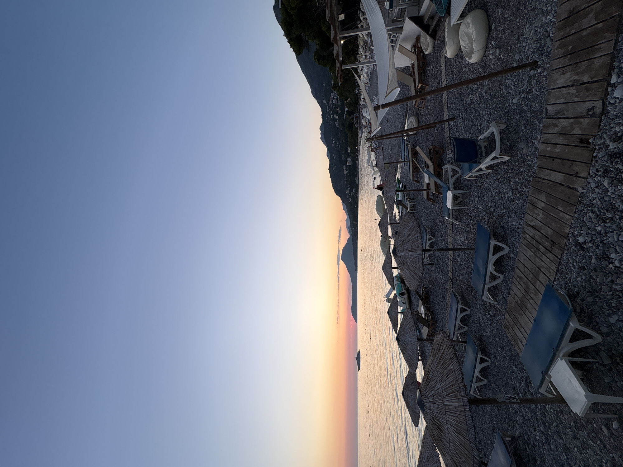 The image shows a pebble beach in the city of Bar, Montenegro, at dusk. The sky is a gradient of pale blue and soft orange, indicating the sun has recently set. A calm sea stretches to the horizon, where a distant boat is visible. The beach is lined with rows of straw umbrellas and empty lounge chairs, suggesting a quiet end to the day. A wooden walkway curves through the beach. In the background, a gentle slope of hills covered in greenery and scattered buildings rises towards the sky. The overall atmosphere is peaceful and serene.