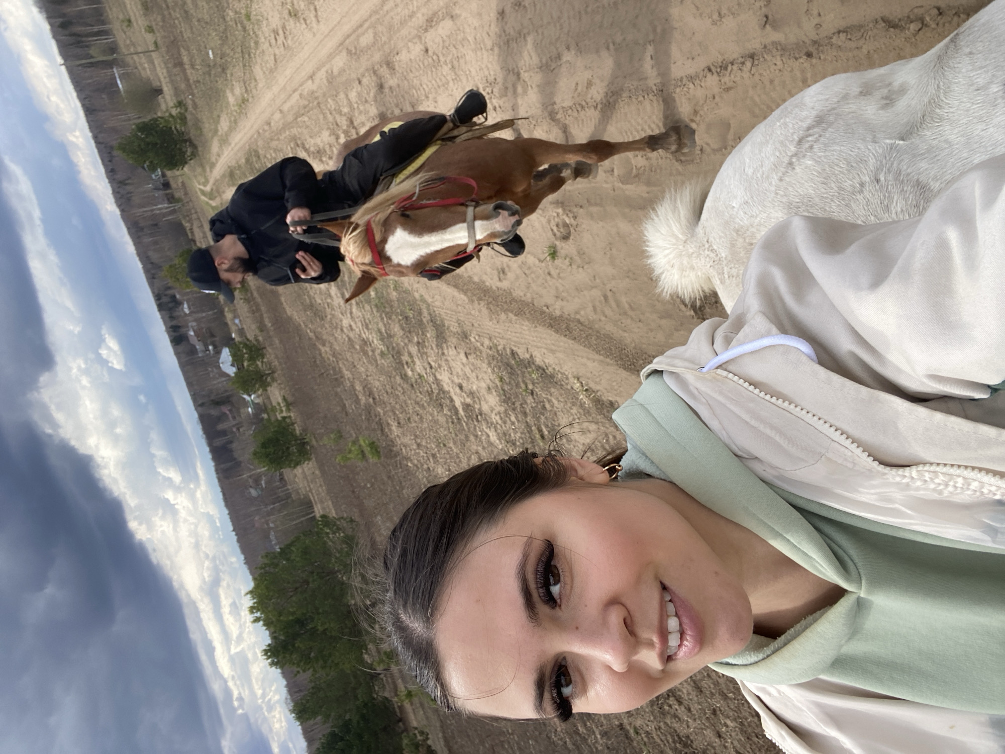 The image captures a woman taking a selfie while horseback riding. She is wearing a light green hoodie and a light-colored jacket. The background features a man on a brown horse, and the overall setting appears to be a sandy field with trees and buildings in the distance. The location is Zhigalki, Russia.