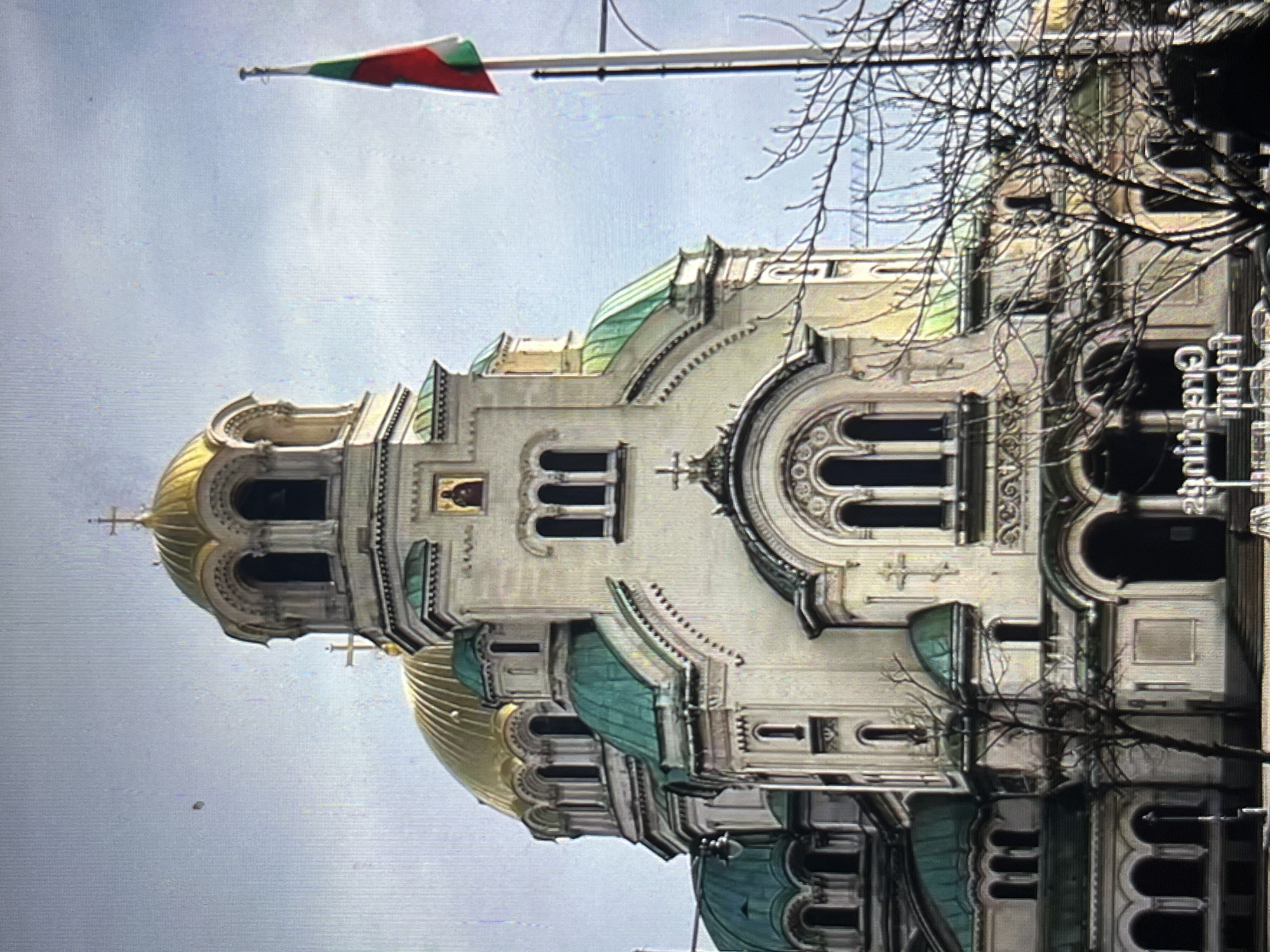 The image depicts the St. Alexander Nevsky Cathedral in Sofia, Bulgaria. The cathedral has a white facade with gold and green domes and is adorned with crosses and decorative architectural details. A Bulgarian flag is visible at the top of the frame. Bare tree branches are seen in the right side of the image. The sky is overcast. There is no indication that the picture was taken in the city of Tinaquillo, Venezuela.
