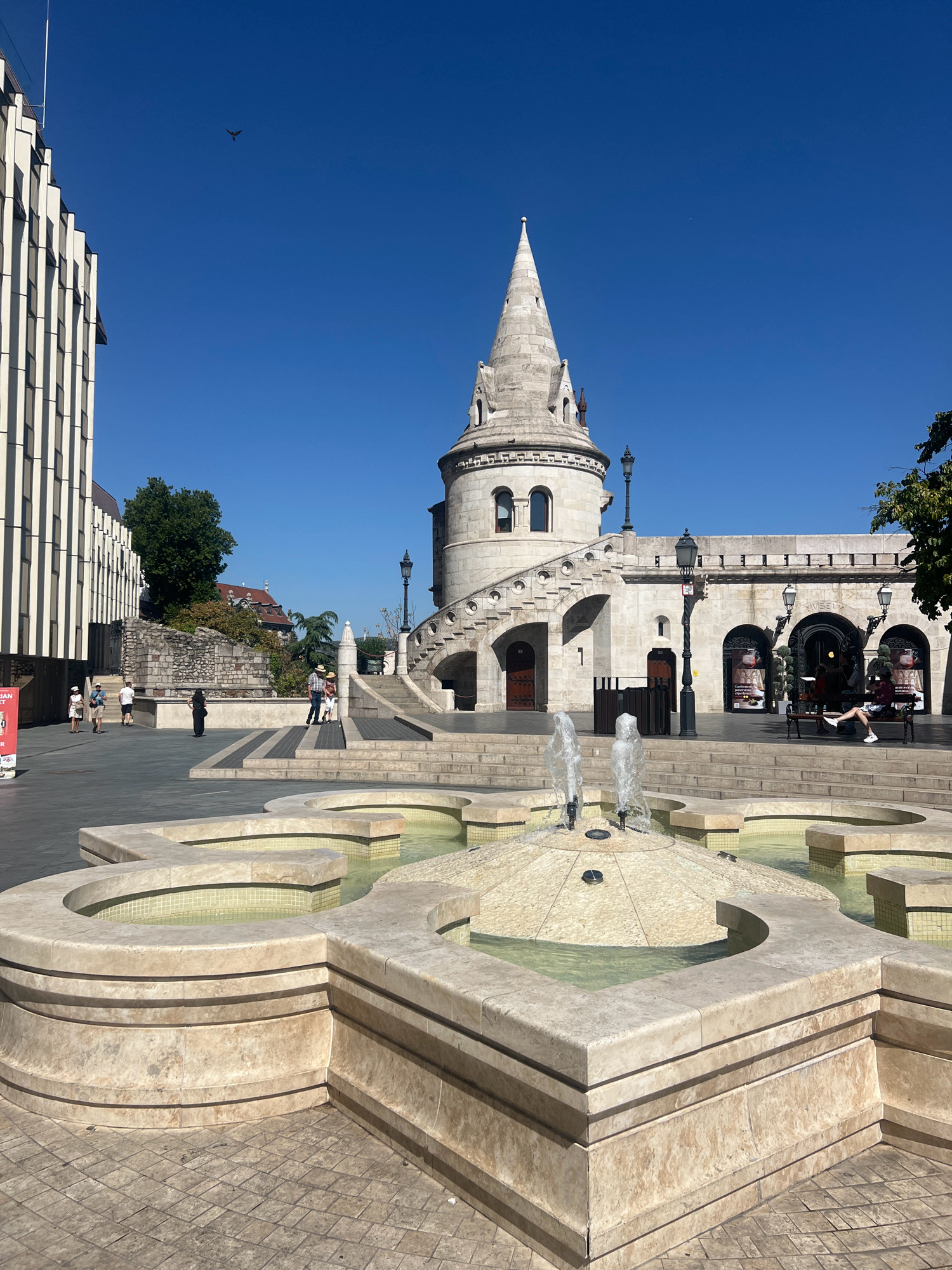 The image shows a view of the Fisherman's Bastion in Budapest, Hungary, on a clear day with a bright blue sky. In the foreground, there is a multi-tiered, geometric fountain made of light-colored stone, with water spraying upwards from its center. Behind the fountain, a wide stone staircase leads up to the Fisherman's Bastion, a Neo-Gothic and Neo-Romanesque structure with turrets and arched walkways. To the left of the staircase, a modern building with a striped facade stands out. Several people are visible walking and standing on the plaza and on the stairs. One person is seated on a bench under an archway of the Bastion. The overall scene depicts a public space with historical architecture and a water feature, popular with tourists.