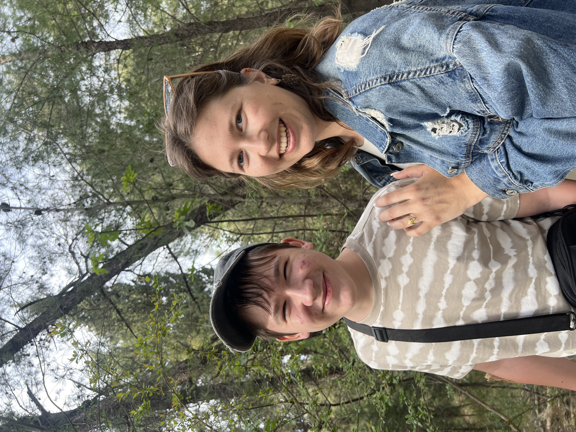 Two people, a woman and a young man, are standing close together in a wooded area, likely a park or forest based on the presence of trees and foliage in the background. The woman is wearing a denim jacket and has sunglasses perched on her head. She is smiling and has her hand resting on the young man's shoulder. The young man is wearing a baseball cap and a patterned t-shirt, and he is also smiling. The setting could potentially be in or near Podgorica, Montenegro, given the environmental context.