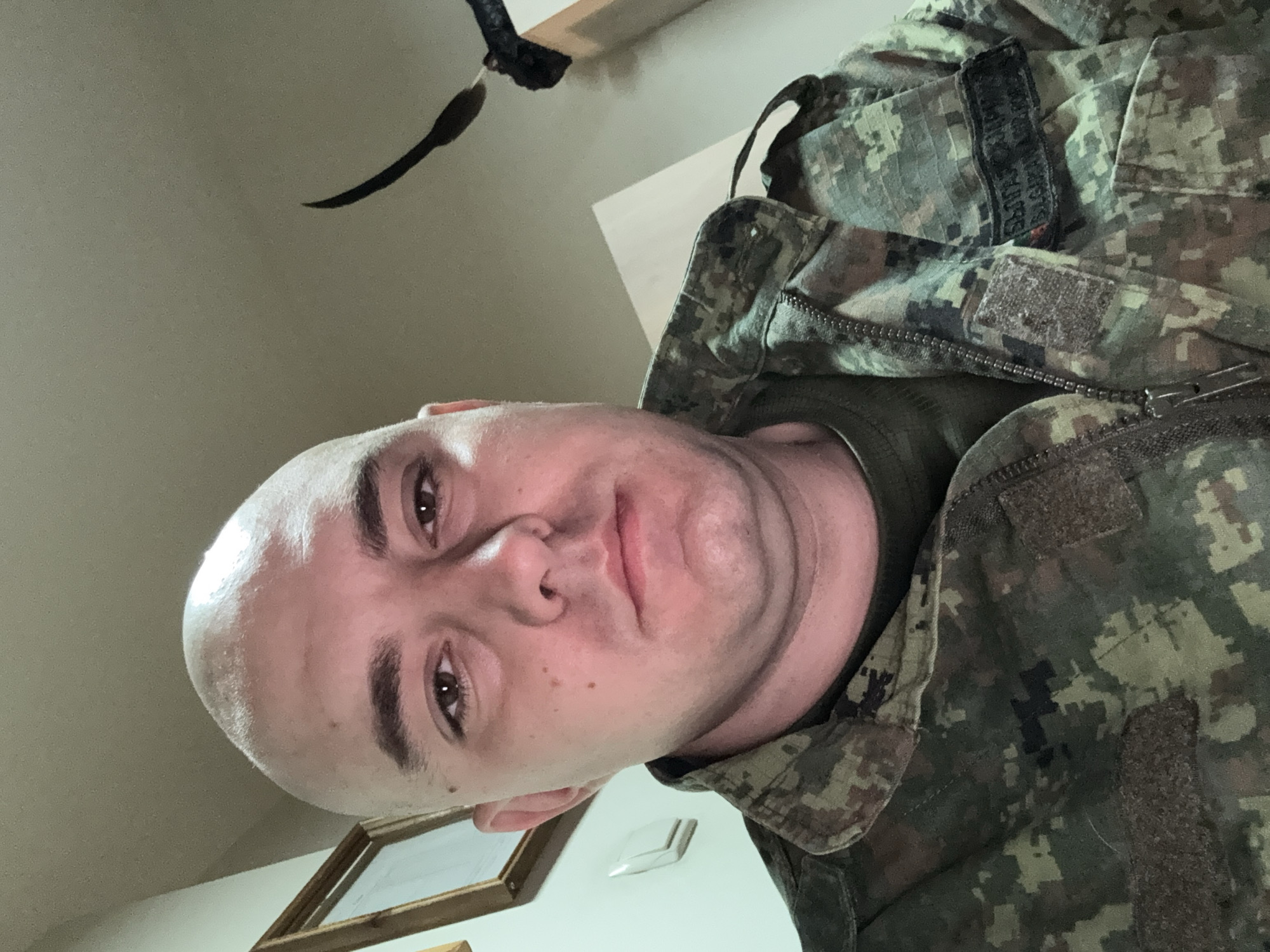 A close-up, slightly angled, indoor portrait shows a bald man wearing camouflage clothing. The camouflage pattern is predominantly green and brown. The man's face is pale, and he has light brown eyes. His lips are full. The clothing has a zipper. On the top of the jacket there appears to be a patch. In the background, blurred and indistinct, are parts of the room, including an object resembling a black bird or decoration hanging from the ceiling, a picture frame on the wall, and a light switch plate. The location is not identifiable as Sofia, Bulgaria based solely on the image.