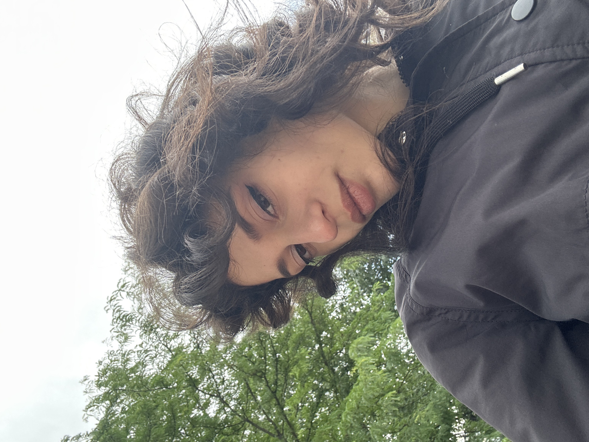 A young woman with dark, curly hair and light skin is captured in a close-up, downward-facing selfie. She is wearing a black jacket with a drawstring closure and metal snaps. Her makeup includes winged eyeliner and a neutral lip color. In the background, lush green foliage from trees and an overcast sky are visible. The image appears to have been taken outdoors, possibly in a park or a residential area with abundant greenery, which could be a characteristic of certain neighborhoods in Sofia, Bulgaria.