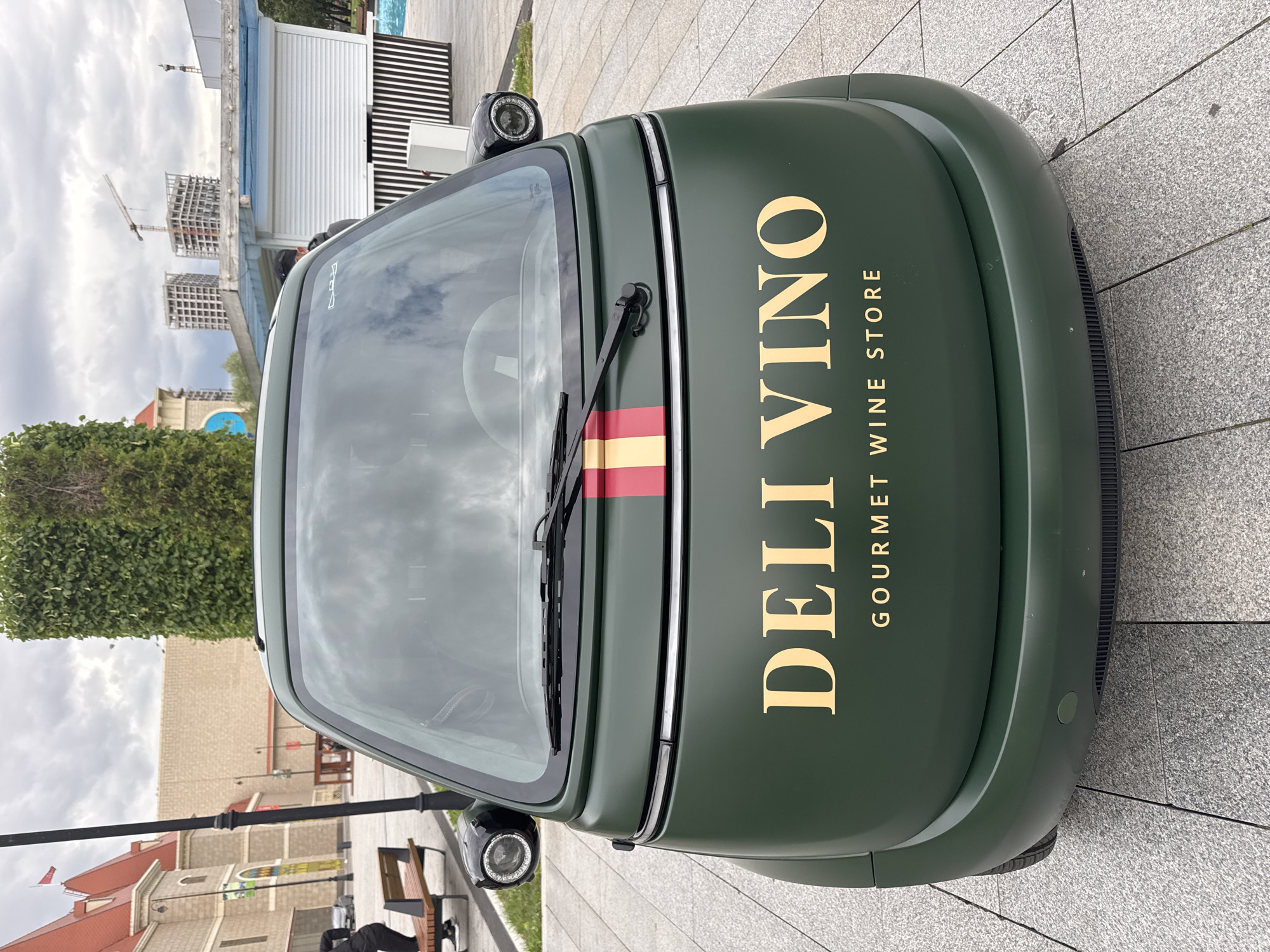 A dark green, three-wheeled vehicle branded with "DELI VINO GOURMET WINE STORE" is parked on a light gray paved surface. The text on the vehicle is in a golden, serif font. A red and yellow stripe is visible across the front of the vehicle, just below the windshield. The vehicle has two headlights on the front, with one clearly visible and the other partially obscured. The scene is set outdoors, with a cloudy sky suggesting it might be daytime. In the background, there are buildings under construction, including a skeleton of a high-rise building and a crane. There are also some green bushes and trees visible. The paving stones are laid in a grid pattern. The overall impression is of an urban setting, possibly a street or a plaza, with some commercial or residential development in progress. The vehicle's branding suggests it is a mobile wine shop. No people are clearly visible interacting with the vehicle.