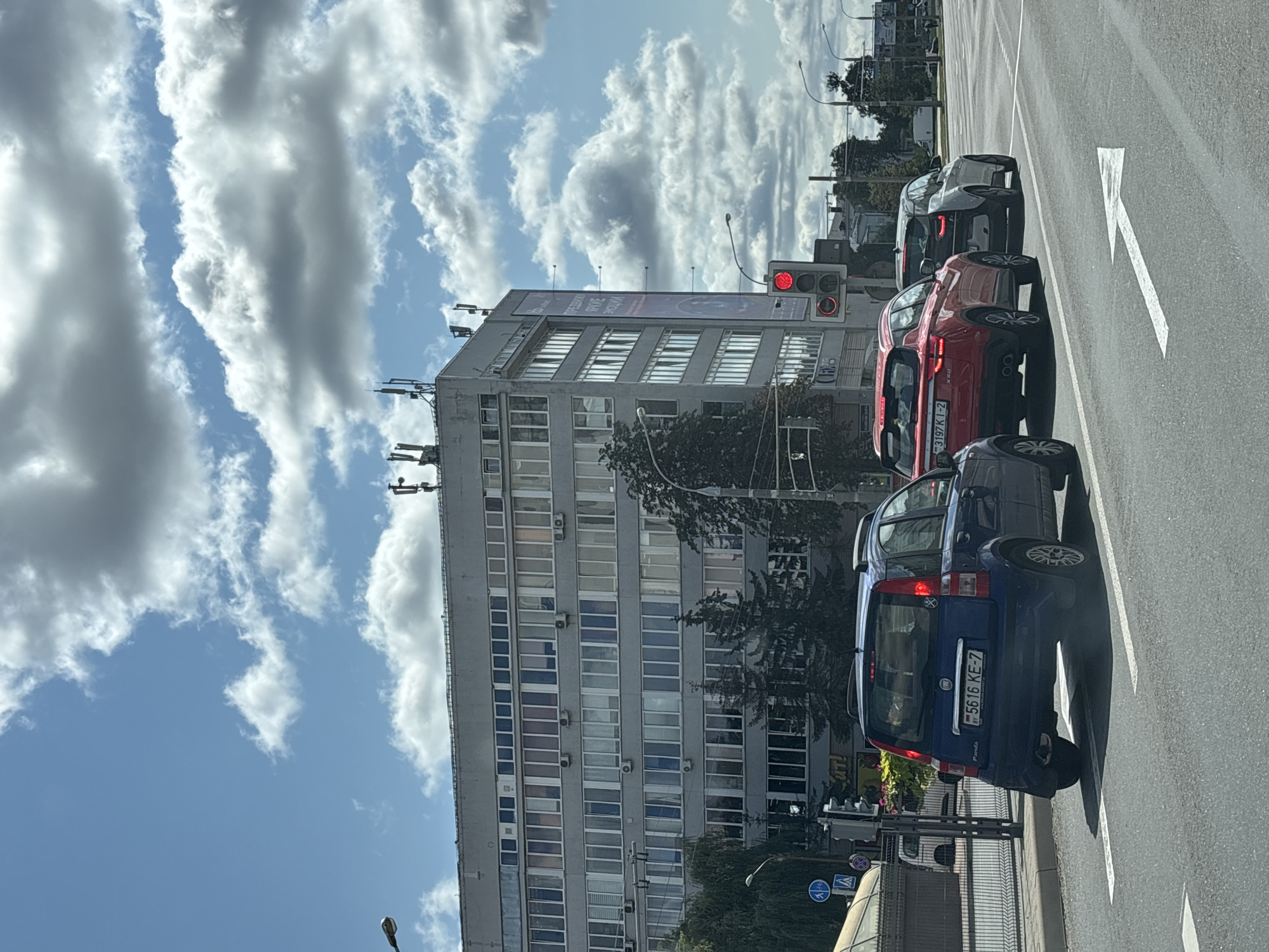 The image captures a street scene in Minsk, Belarus, likely in a commercial or administrative district, given the large, multi-story building dominating the background. The building, constructed of light-colored concrete or stone, features a grid of uniformly spaced windows. On its roof, several communication antennas are visible. In front of the building, a multi-lane road is depicted, with several vehicles in traffic. The foreground shows the rear of a blue Fiat Panda with license plate "BY 5616 KE-7". To its right is a red SUV, possibly a Geely, with license plate "3197 K-2". Further back, a white car, possibly an electric vehicle given its design, is also in traffic. A traffic light in the middle ground is illuminated with a red signal, indicating that the vehicles are stopped. The sky is partly cloudy, with fluffy white clouds against a blue backdrop. Trees are visible along the side of the road and near the building. The overall impression is a typical urban environment on a bright, partly cloudy day.