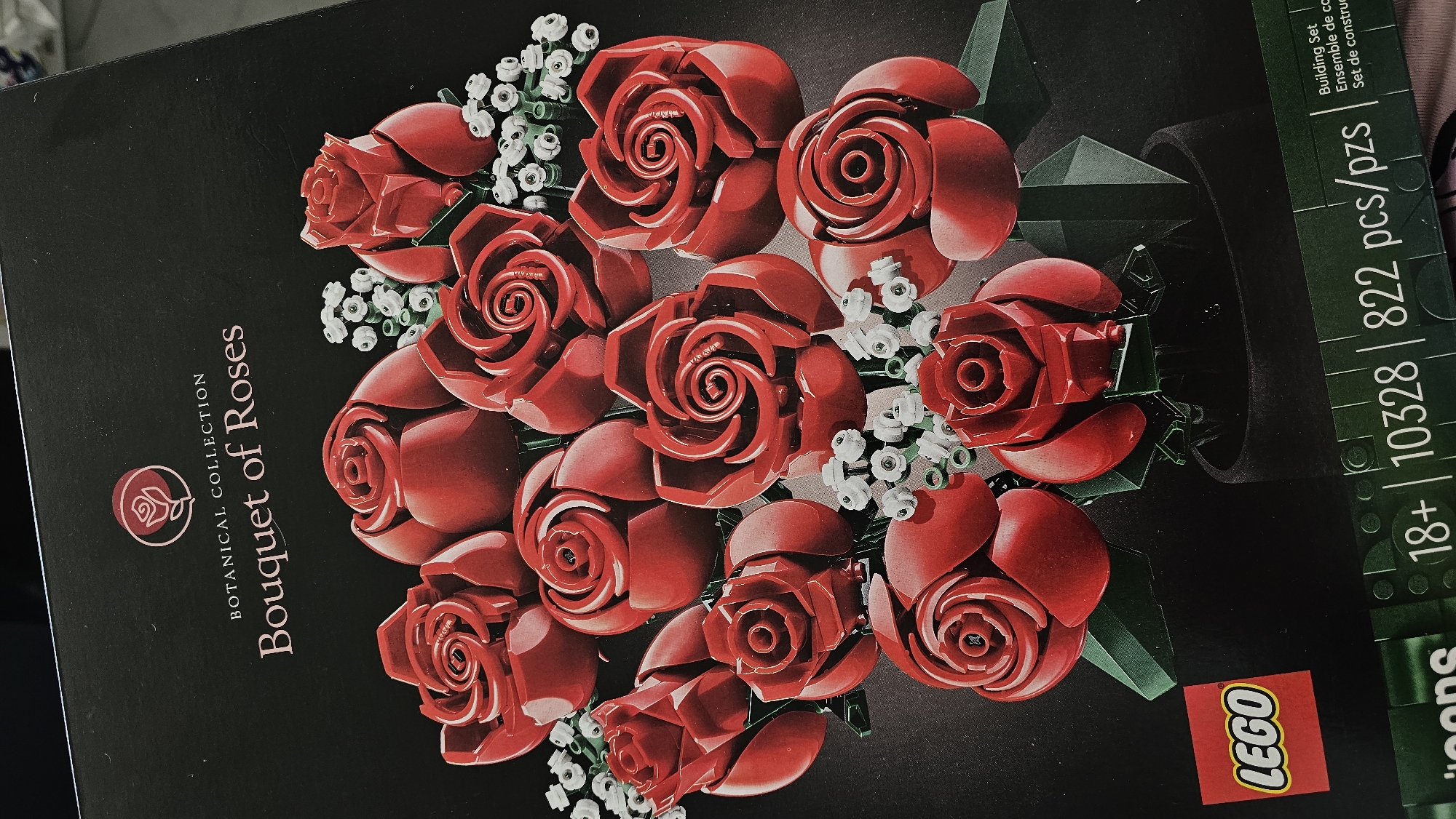 The image shows the box of a LEGO Botanical Collection Bouquet of Roses set. The box is primarily black, and the main image is of a bouquet of red LEGO roses with white LEGO baby's breath flowers. Below the bouquet, on the right side of the box, is the LEGO logo in red and white. Above this is the "18+" age recommendation, the set number "10328", and the number of pieces "822 pcs/pzs". The text "BOTANICAL COLLECTION" and "Bouquet of Roses" is printed in white to the left of the roses. The image does not contain any indications that it was taken in Puebla, Mexico.