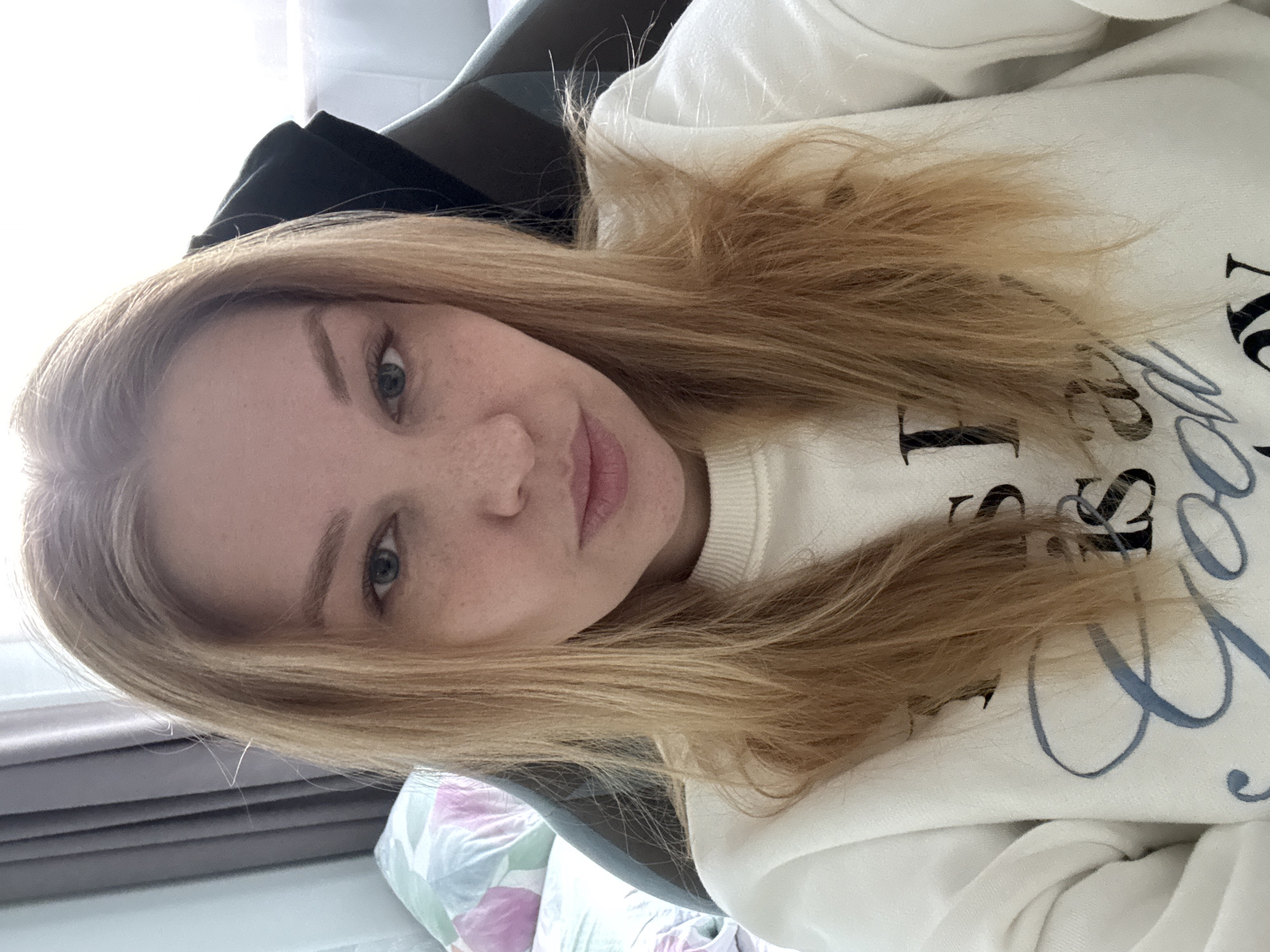 A selfie photograph of a young, light-skinned woman with blonde hair and freckles. She is wearing a white sweatshirt with black and blue lettering. The background includes a dark chair and a glimpse of a window with light curtains. The woman is looking directly at the camera. There is no evidence that this image was taken in the city of Rusanovo, Russia.