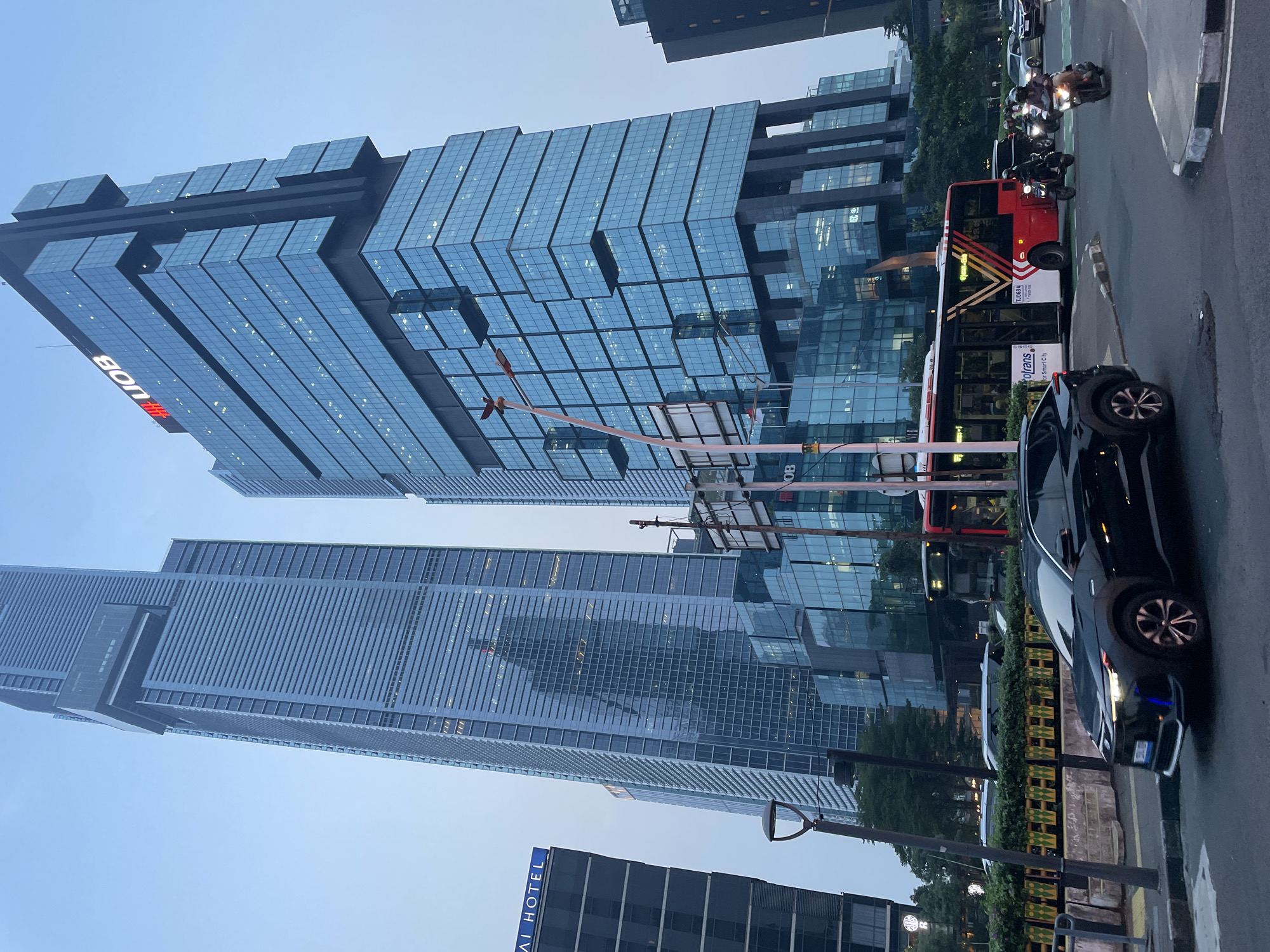 The image presents a vibrant urban street scene in Central Jakarta, Indonesia, captured during what appears to be late afternoon or early evening. The perspective is looking upwards, showcasing a series of modern skyscrapers that define the city's skyline.
Dominating the center is a striking UOB tower, characterized by its distinctive blue glass facade arranged in stepped, zig-zagging panels that reflect the pale blue sky. The prominent red "UOB" logo is visible on its upper section. To its left, another tall, contemporary building rises, featuring a facade of uniform glass and steel panels. In the lower left foreground, a darker, multi-story building partially displays the text "AI HOTEL."
At street level, the scene is busy with traffic. A red public transit bus, identifiable by its "oltrans" and "Smart City" branding along with the number "T20694," is visible on the right side of the road, suggesting it's part of Jakarta's TransJakarta system. A black car is either parked or stopped in front of the bus, and further right, several motorcycles with riders are present. The street features a painted green lane, likely for dedicated bus or bike traffic, and is lined with curbs, street poles, and a decorative barrier with yellow and green panels. Some internal lights are visible within the buildings, indicating the transition from day to evening in this bustling metropolitan area.