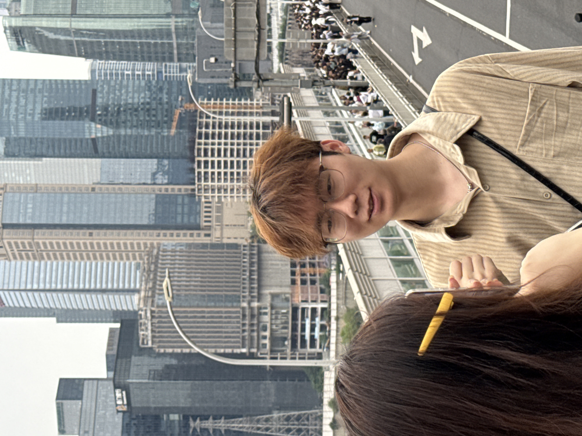 The image depicts a man with brown hair and glasses standing on a bridge in Yuzhong District, China. Behind him, high-rise buildings form the urban landscape. A crowd of people is visible on the bridge, partially obscured. A woman with dark hair is in the foreground, partially obscuring the view. The man is wearing a tan striped shirt and a silver necklace. He is holding a cell phone. The environment suggests a modern, urban setting.