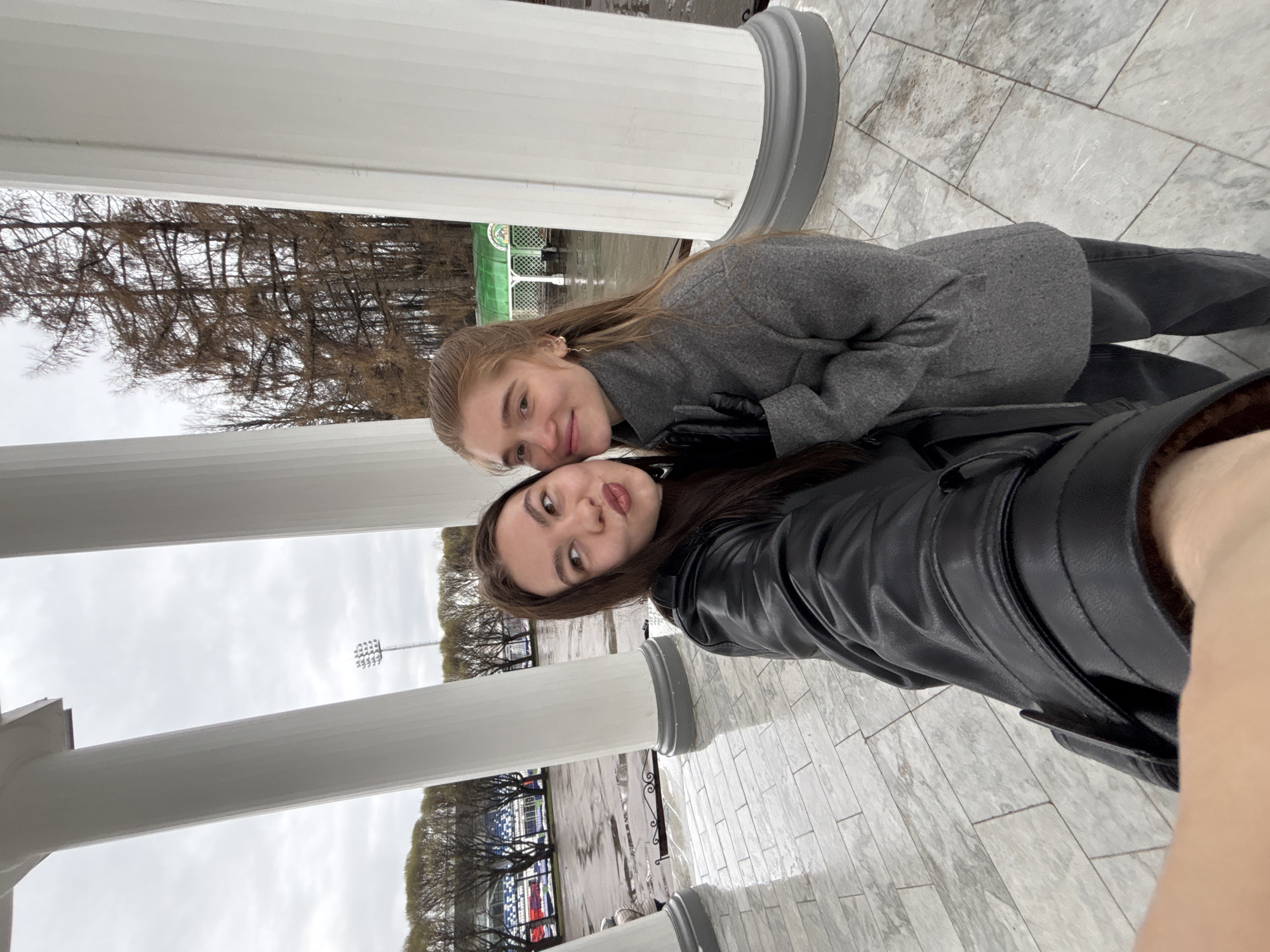 Two women are posing for a selfie in Perm, Russia. They are near white, classical-style columns. The woman in the foreground wears a black leather jacket, and the woman behind her wears a gray wool coat. They are both wearing black gloves. Behind them is a view of a city street with trees and buildings. A light pole and a building resembling a stadium can be seen. The pavement is tiled, and it appears to be damp or recently rained upon.