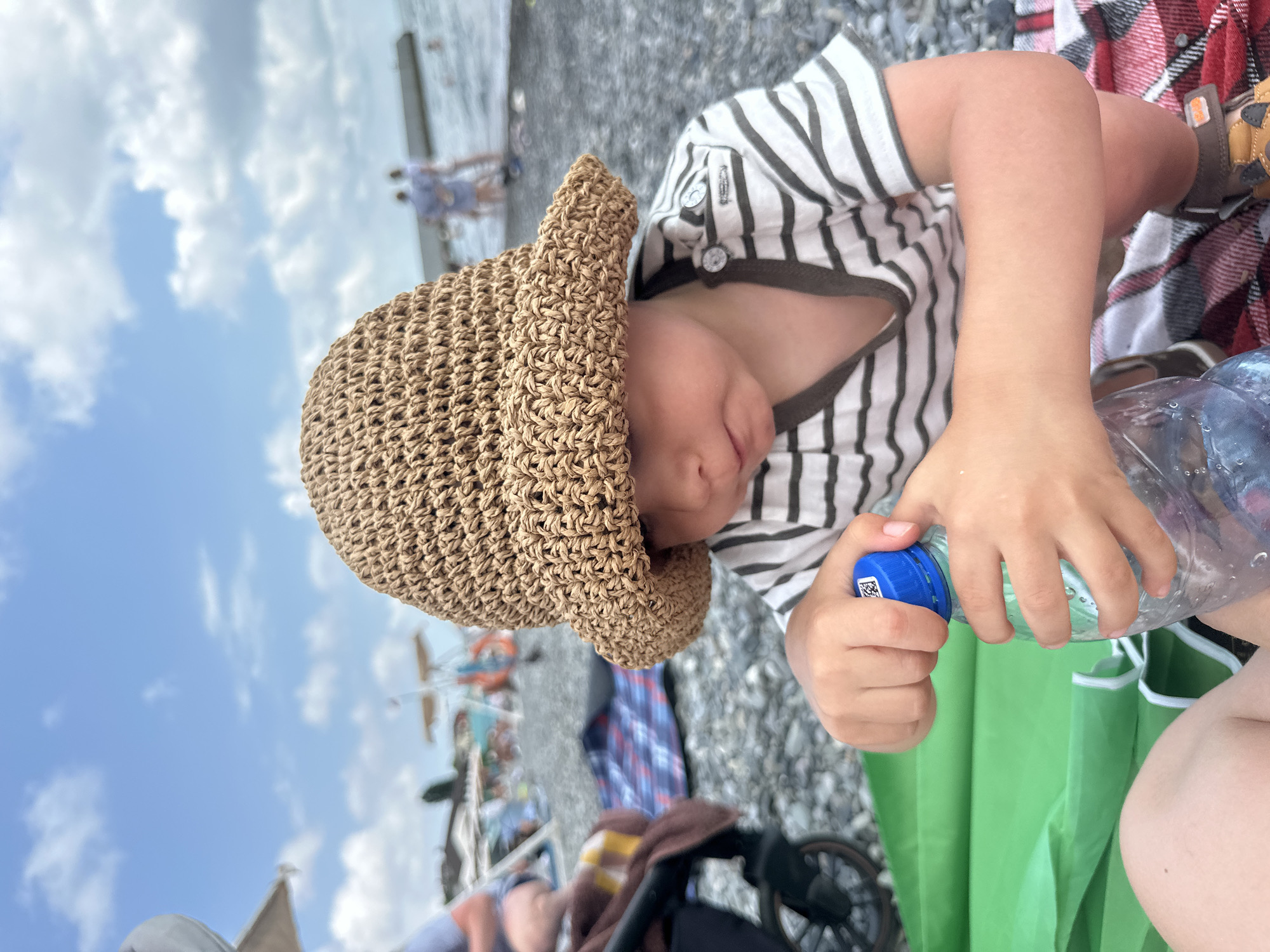 A child wearing a striped t-shirt and a straw hat is holding a clear plastic bottle with a blue cap, appearing to pour water from it. The child's face is partially obscured by the hat, but their hands are actively engaged with the bottle. They are seated on a beach with pebbles and what looks like a checkered blanket nearby. In the background, there are blurry figures of people and some structures, suggesting a public beach or boardwalk area. The sky is visible, with some clouds. The overall scene suggests a casual outdoor activity, likely on a sunny day, given the presence of a hat and the bright sky. There is some text visible on the blue cap of the bottle, but it is too pixelated to read. The image is taken from a slightly low angle, looking up towards the child.