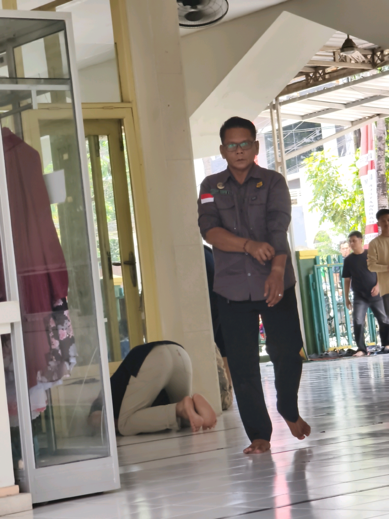 In an outdoor covered walkway, possibly a mosque in Samarinda, Indonesia, a man in a dark grey uniform with glasses walks to the right, his left hand adjusting his sleeve. He wears a badge with a flag and a crest, and a bracelet on his left wrist. To his left, a person in a dark shirt and light-colored pants is kneeling on the floor in prayer, their head bowed. A large glass display case stands to the far left. In the background, other individuals are visible, including one in a black t-shirt and two others further back. The ground is tiled, and a green fence runs along the right side of the walkway.