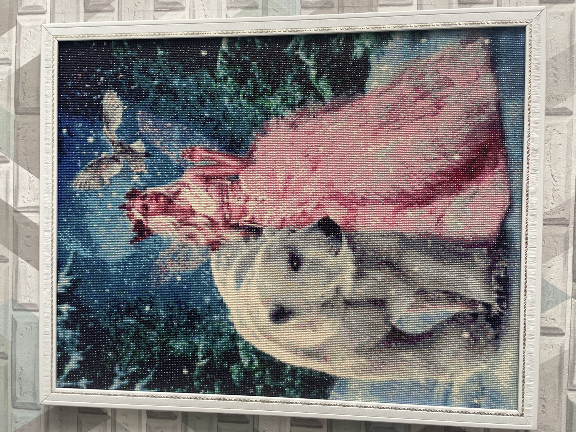 The image is a framed diamond painting of a girl in a pink dress with a panda bear, set against a backdrop of green foliage and a blue sky with a bird. The frame is white and the painting is hanging on a wall with a brick pattern. There is no indication of location in the city of Vladivostok, Russia.