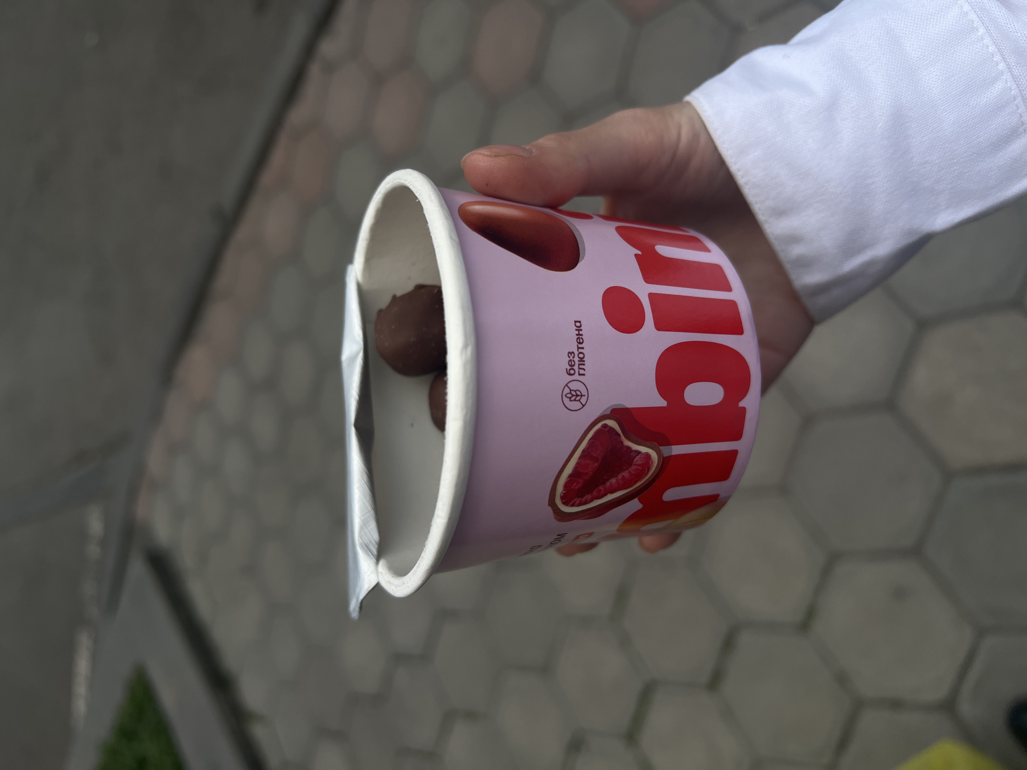 A slightly high-angle, outdoor shot depicts a hand holding a cylindrical container with chocolate inside. The container, primarily pink, is decorated with images of chocolate-covered raspberries. Text in red is also visible on the side of the container. The hand holding the container is wearing a white shirt or lab coat. The background shows a section of a paved area composed of light gray hexagonal stones. The location appears to be an urban or suburban setting.