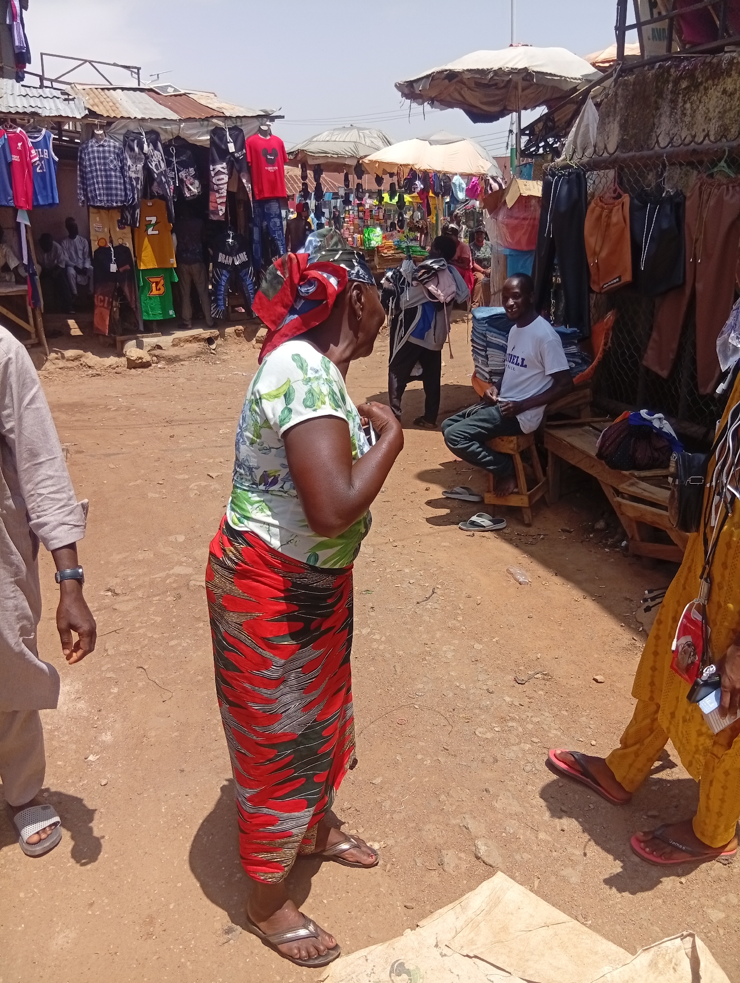 In Bukuru, Nigeria, an outdoor market stall displays a variety of clothing, including shirts, shorts, and pants, on hangers and shelves. The stall is constructed with a corrugated metal roof and wire mesh fencing. A man in a white t-shirt and dark pants sits on a wooden stool, looking towards the camera. Several individuals are browsing or standing around the stall, including a woman wearing a colorful headscarf, a green and white floral-patterned shirt, and a long red and black patterned skirt. Another person, partially visible on the left, wears a light-colored traditional outfit and a dark watch. The ground is unpaved, consisting of dirt and sand. In the background, other market stalls and people are visible, under the shade of umbrellas.