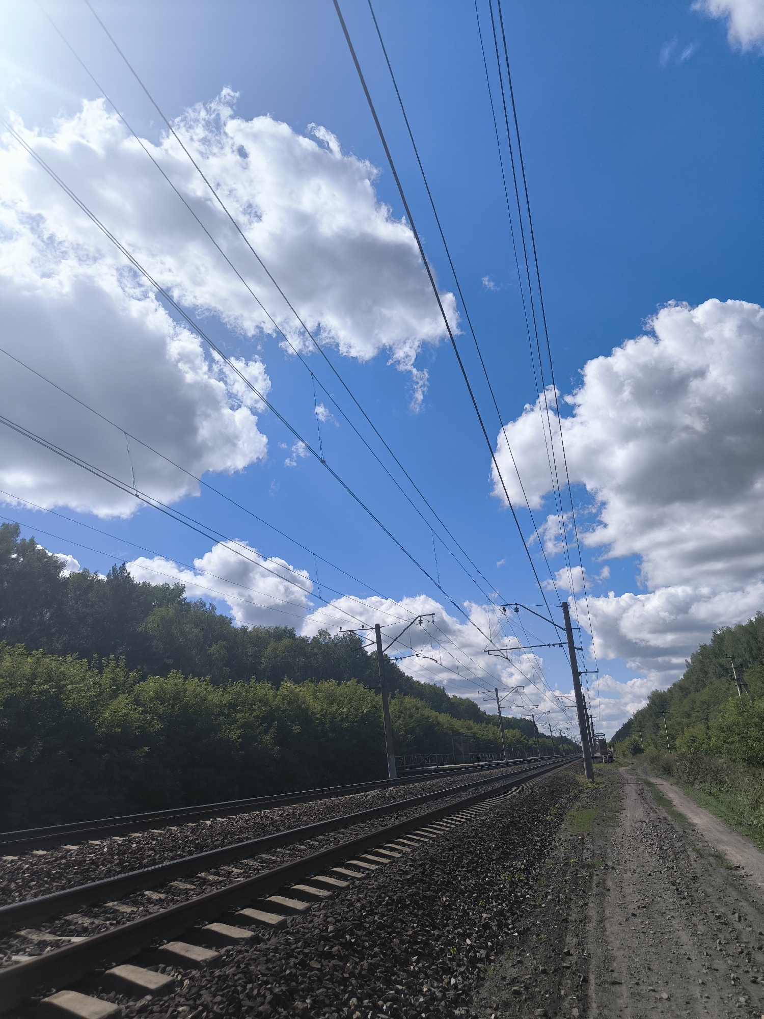 An eye-level shot captures a section of active railroad tracks in Maiskii, Russia, running diagonally from the lower left to the upper right of the frame. The tracks are flanked by a gravelly dirt path on the right and a dense line of lush green trees on the left, which form a natural embankment. Overhead, a vast expanse of blue sky is dotted with large, fluffy white clouds. Numerous power lines, suspended from poles along the route, stretch across the sky, creating a linear pattern that recedes into the distance. The scene appears to be a bright, sunny day, with some lens flare visible in the upper left corner. No people or animals are visible in the image.